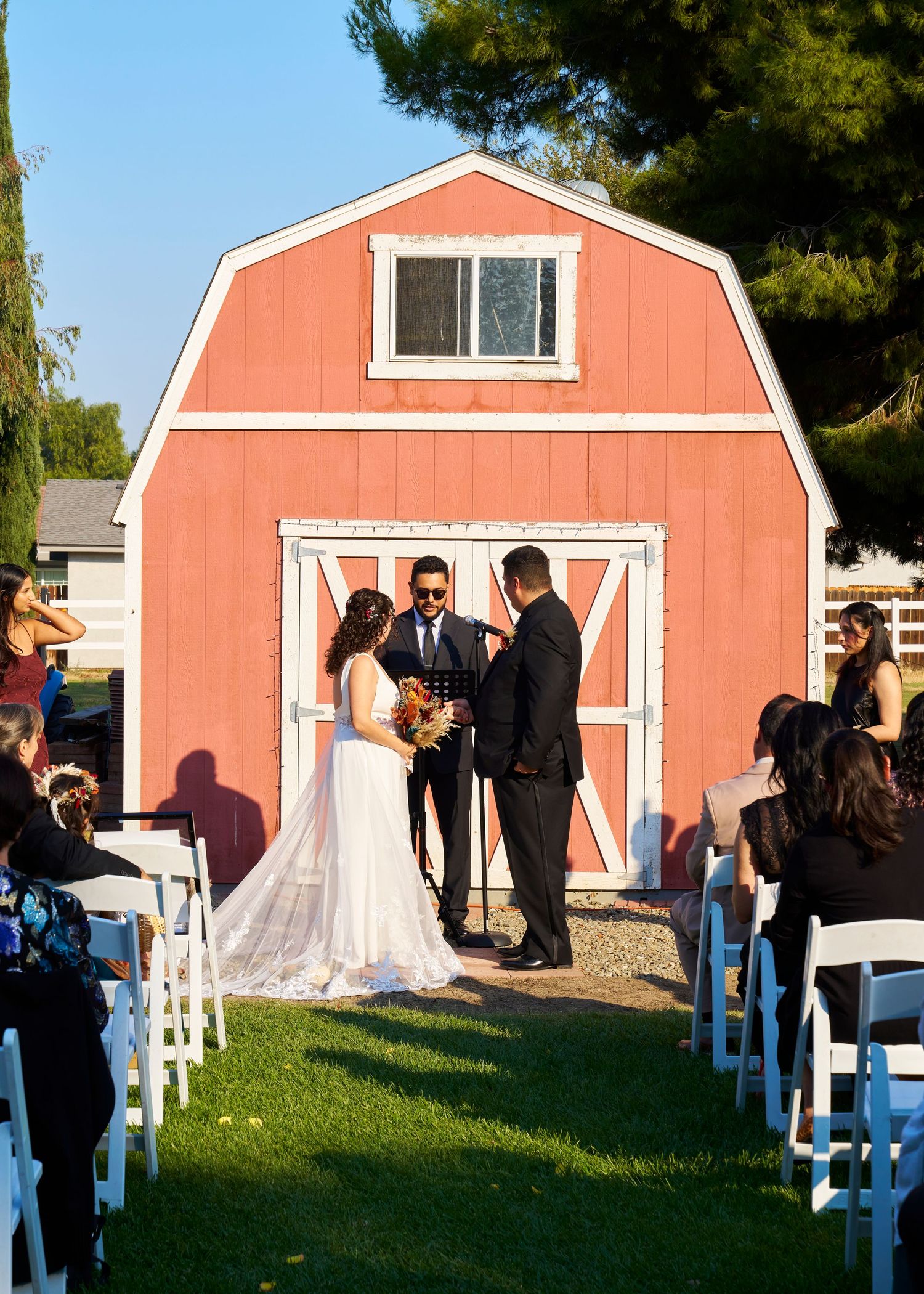 Couple exchanging vows outdoors in front of a charming red barn with white trim during a sunny wedding ceremony.