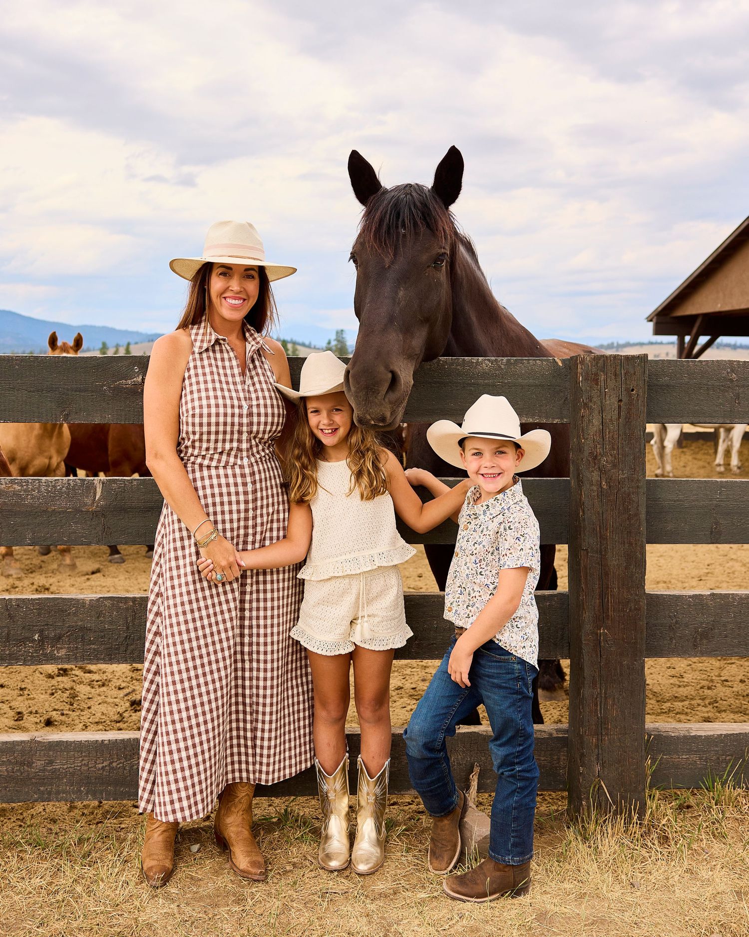 A mother and two children in cowboy hats pose with a dark horse at a wooden ranch fence.