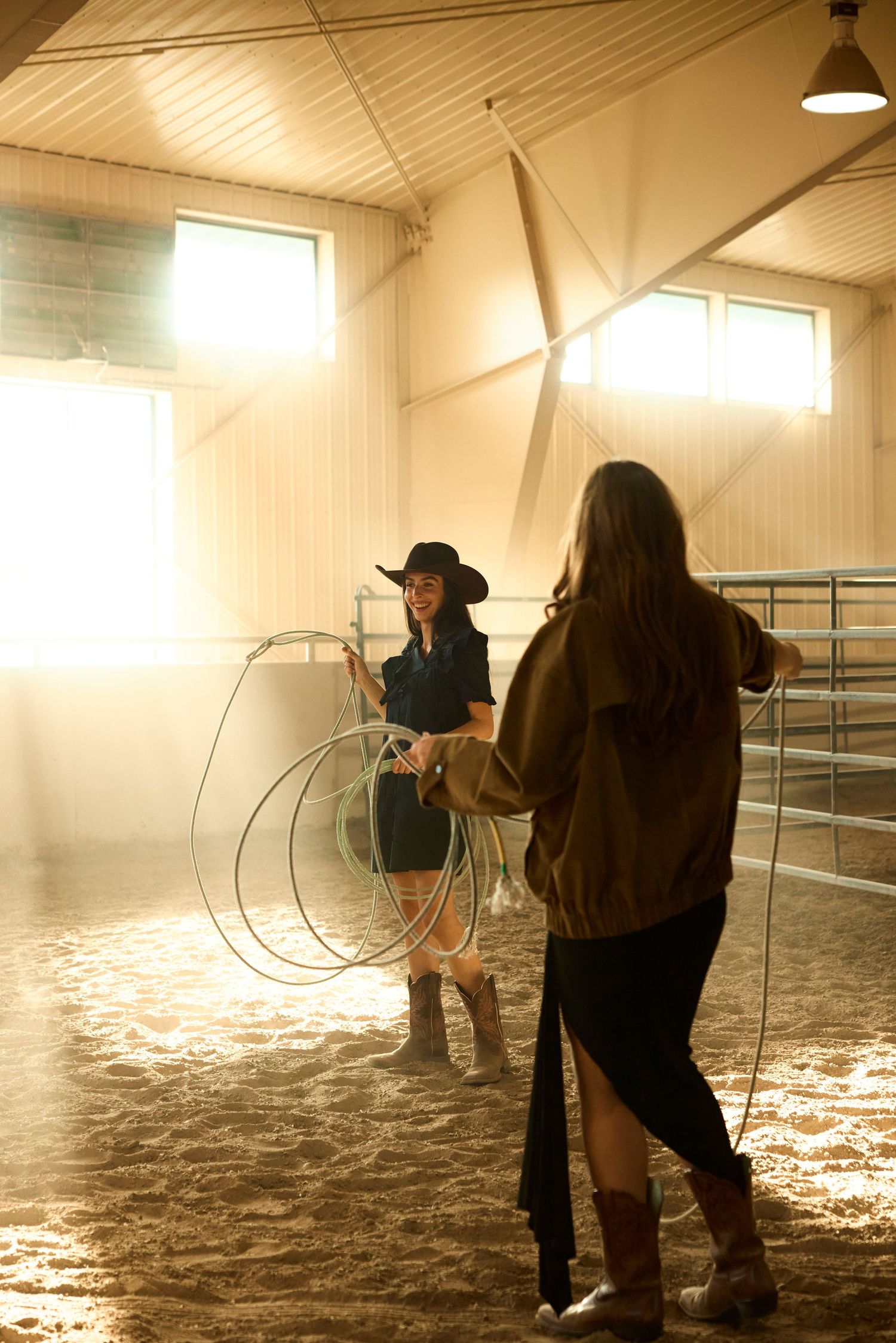 Two women learning to rope with lassos inside a golden sunlit barn arena.