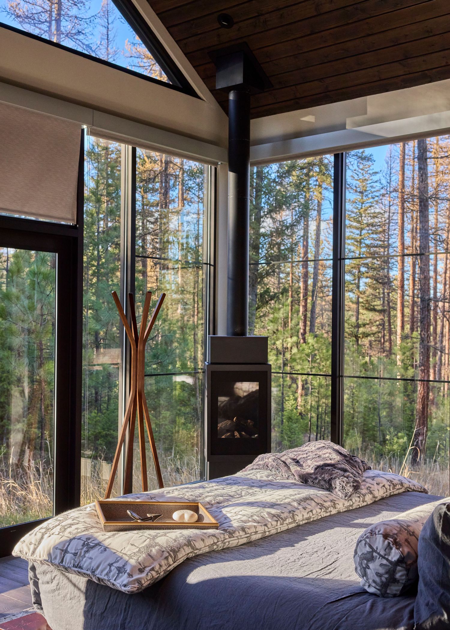 A cozy forest cabin bedroom with floor-to-ceiling windows, a wood-burning fireplace, and a bed with nature views.