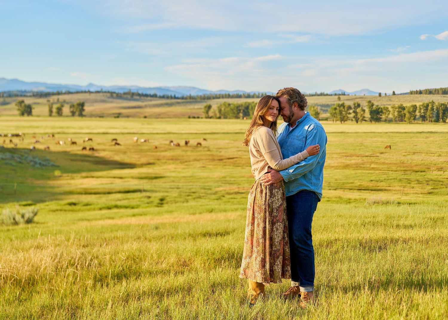 Couple holds each other close in sunlit golden field with scenic mountain valley backdrop.