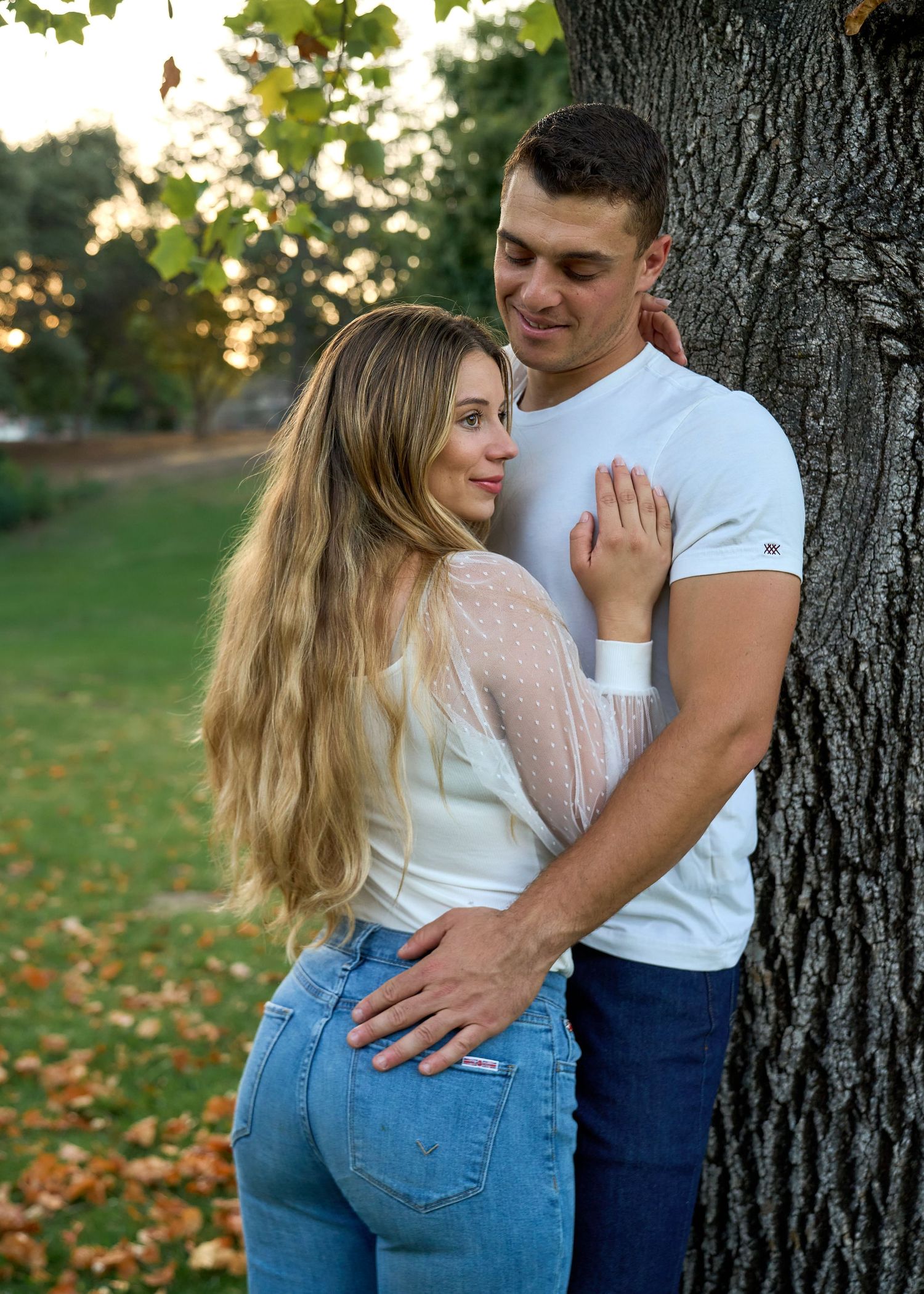 Couple leaning against large tree trunk in autumn park, woman in jeans and lace top.