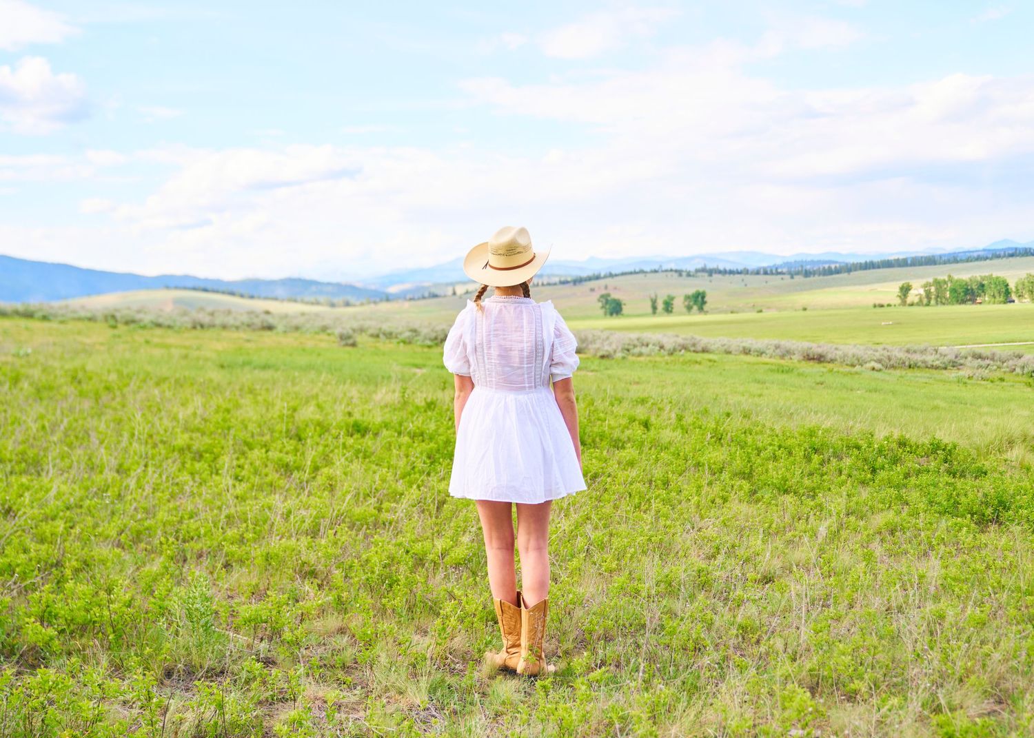 Woman in white dress and straw hat standing with back to camera in open green mountain meadow.