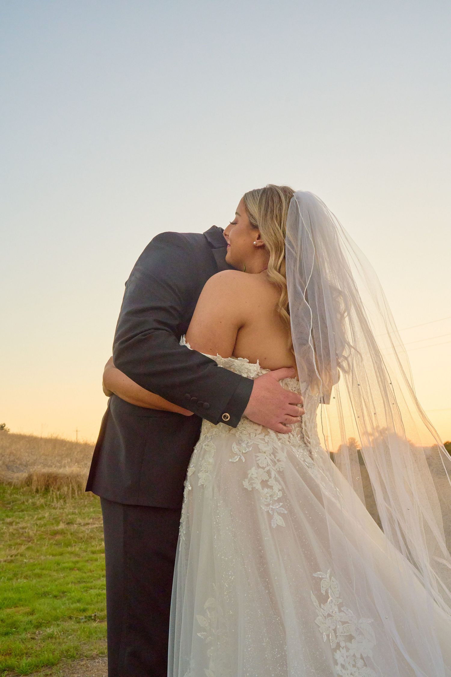 Bride and groom sharing a romantic embrace at sunset, bride wearing a flowing lace gown and long veil in golden light.