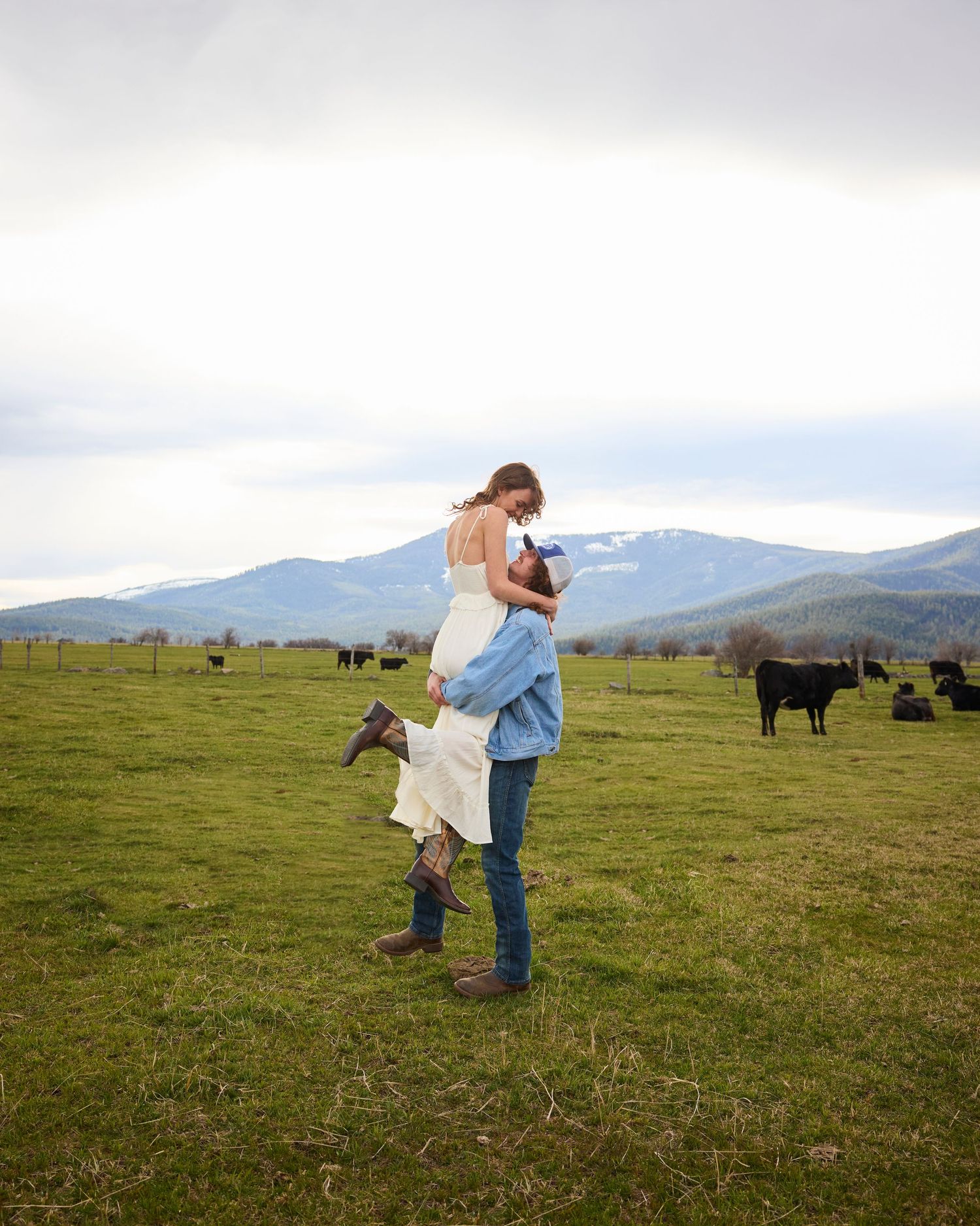 Man lifting woman in white dress and cowboy boots in a green pasture with cattle and mountains.