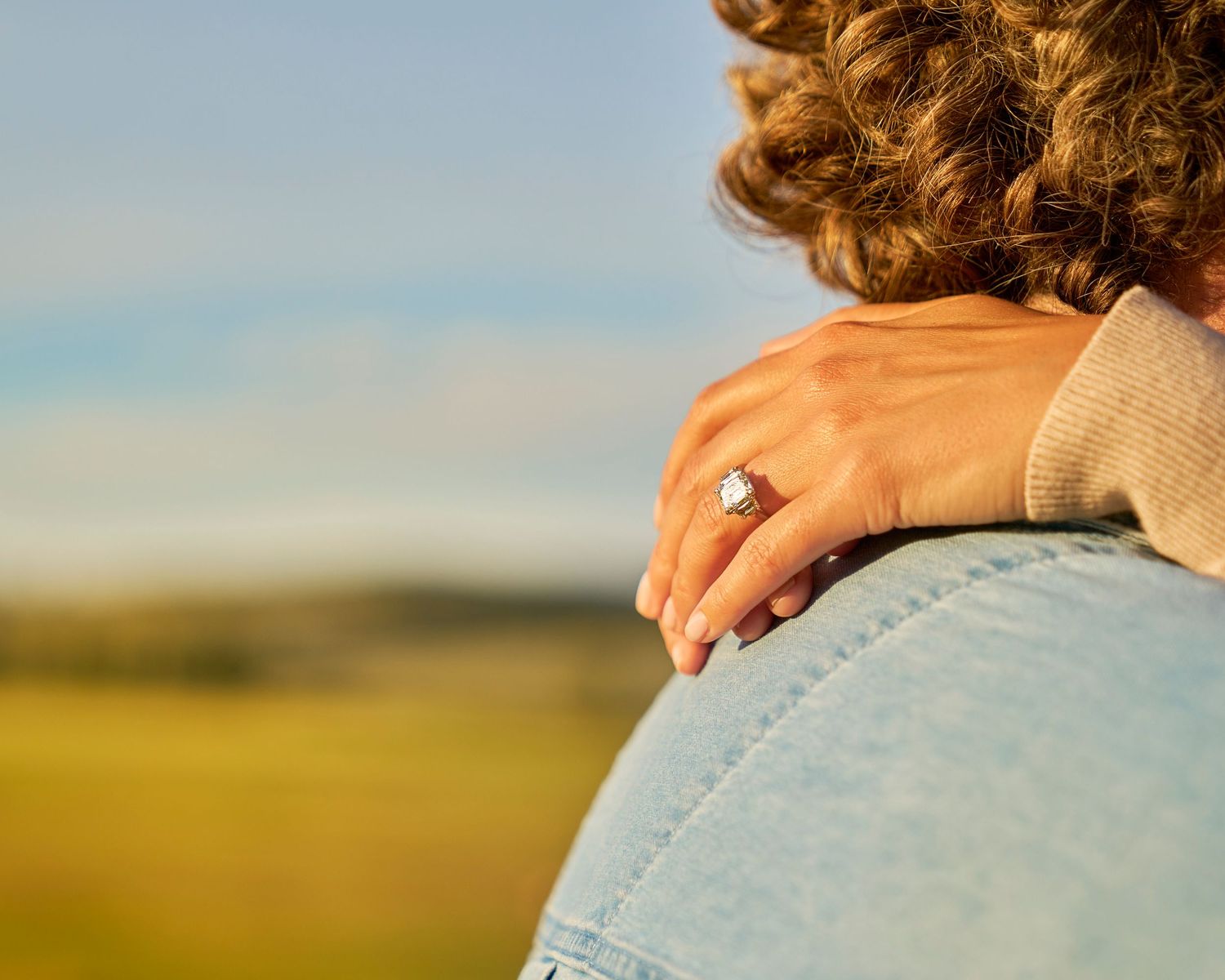 Close-up of woman's hand with diamond engagement ring resting on man's shoulder outdoors.