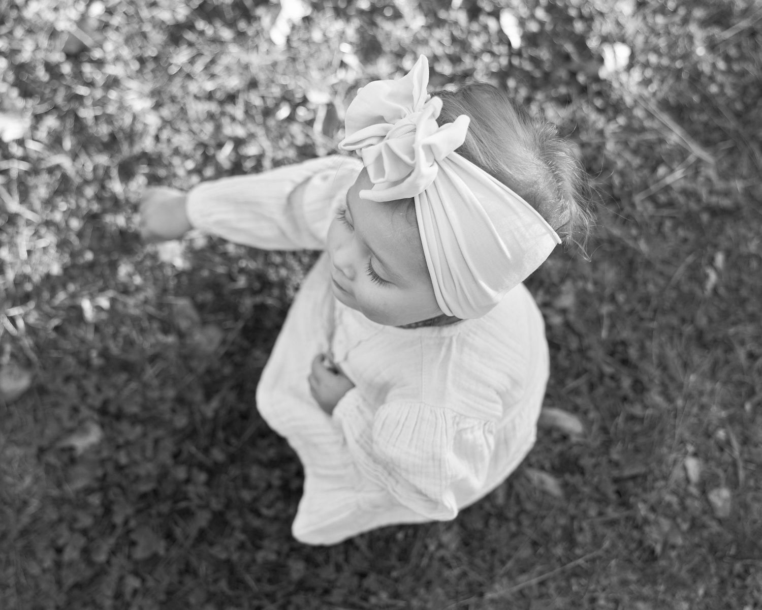 Black and white overhead photo of a baby wearing a bow headband sitting outdoors on the ground.