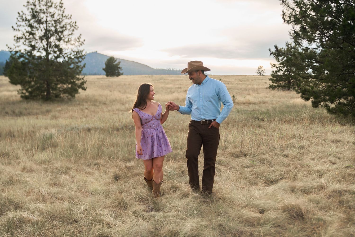 A couple in western wear holds hands while walking through a golden meadow with trees and mountains behind.