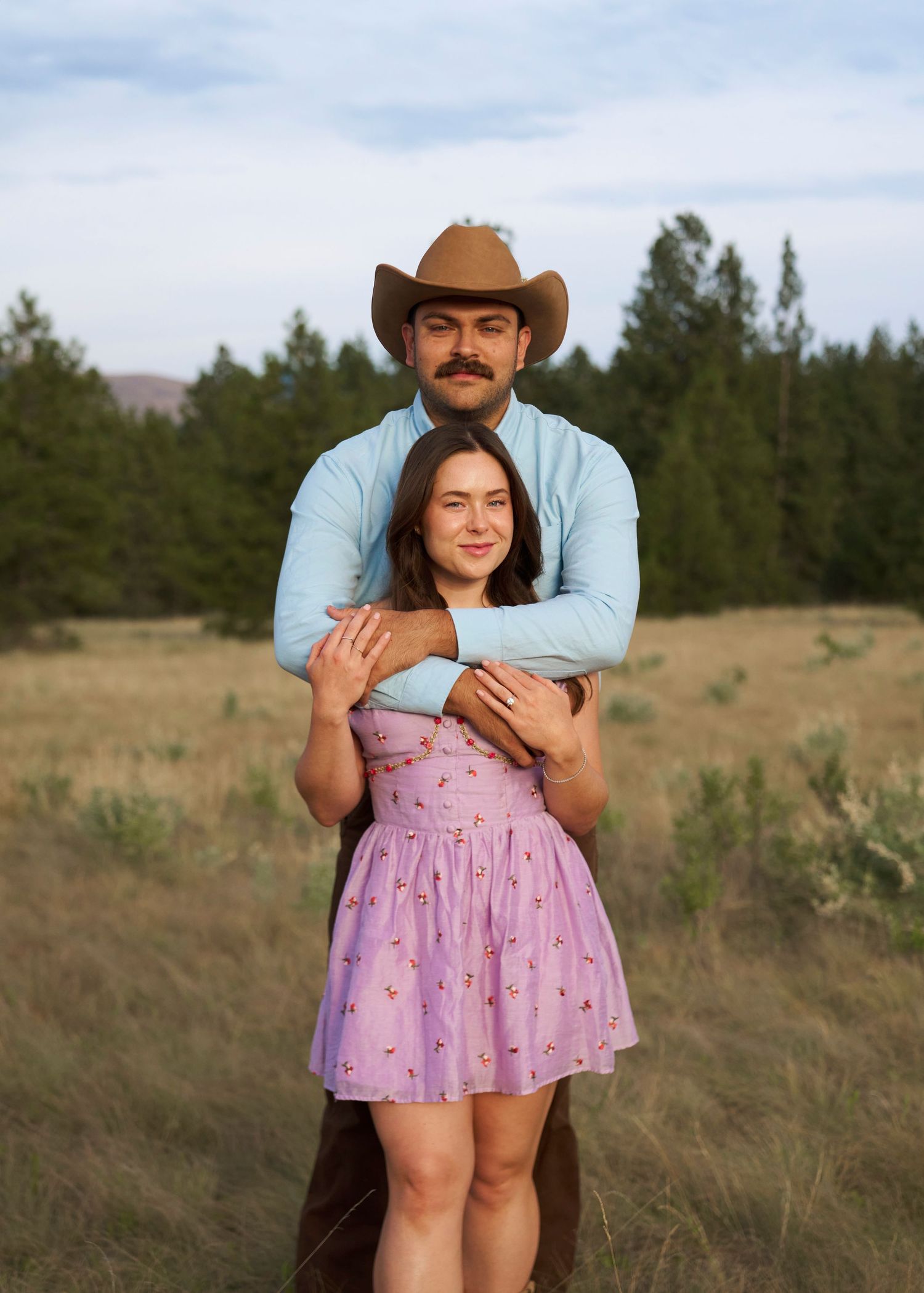 Man in cowboy hat and blue shirt embracing woman in purple floral dress from behind in a rustic meadow setting.