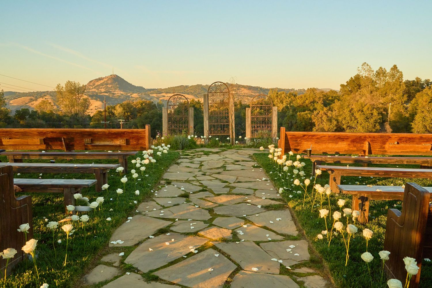 Stone aisle lined with white roses leads to rustic pergola altar with mountain views at sunset ceremony.
