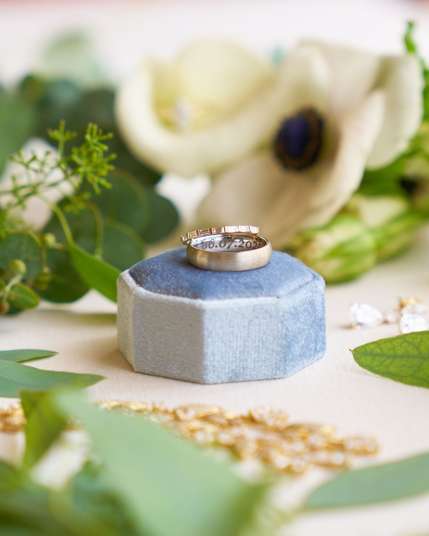 Two wedding bands displayed on a blue velvet hexagonal ring box surrounded by white flowers and greenery.