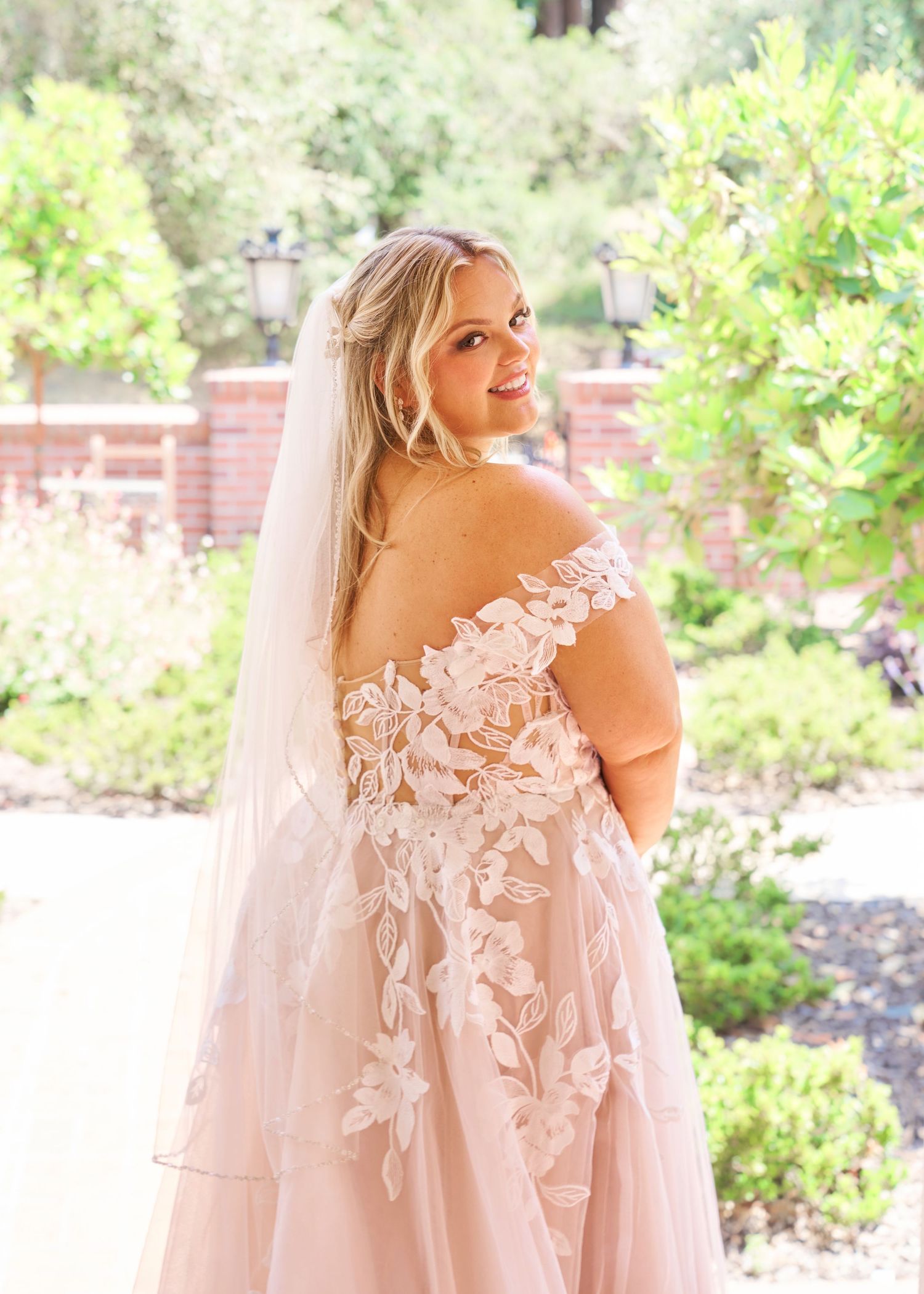 Beautiful bride in off-shoulder floral lace wedding gown with veil posing outdoors in garden.