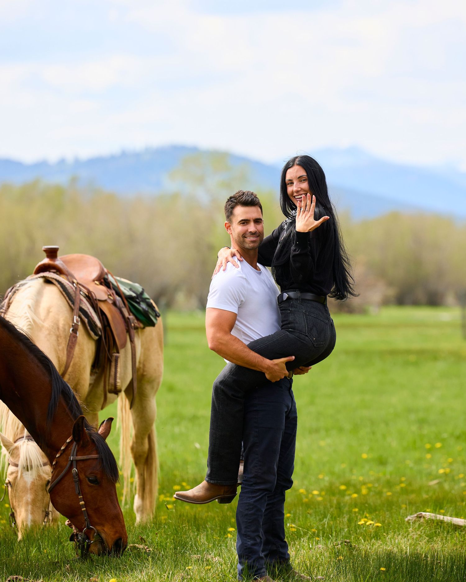 Man lifting woman in a green meadow as she shows off her engagement ring, with a palomino horse grazing nearby.