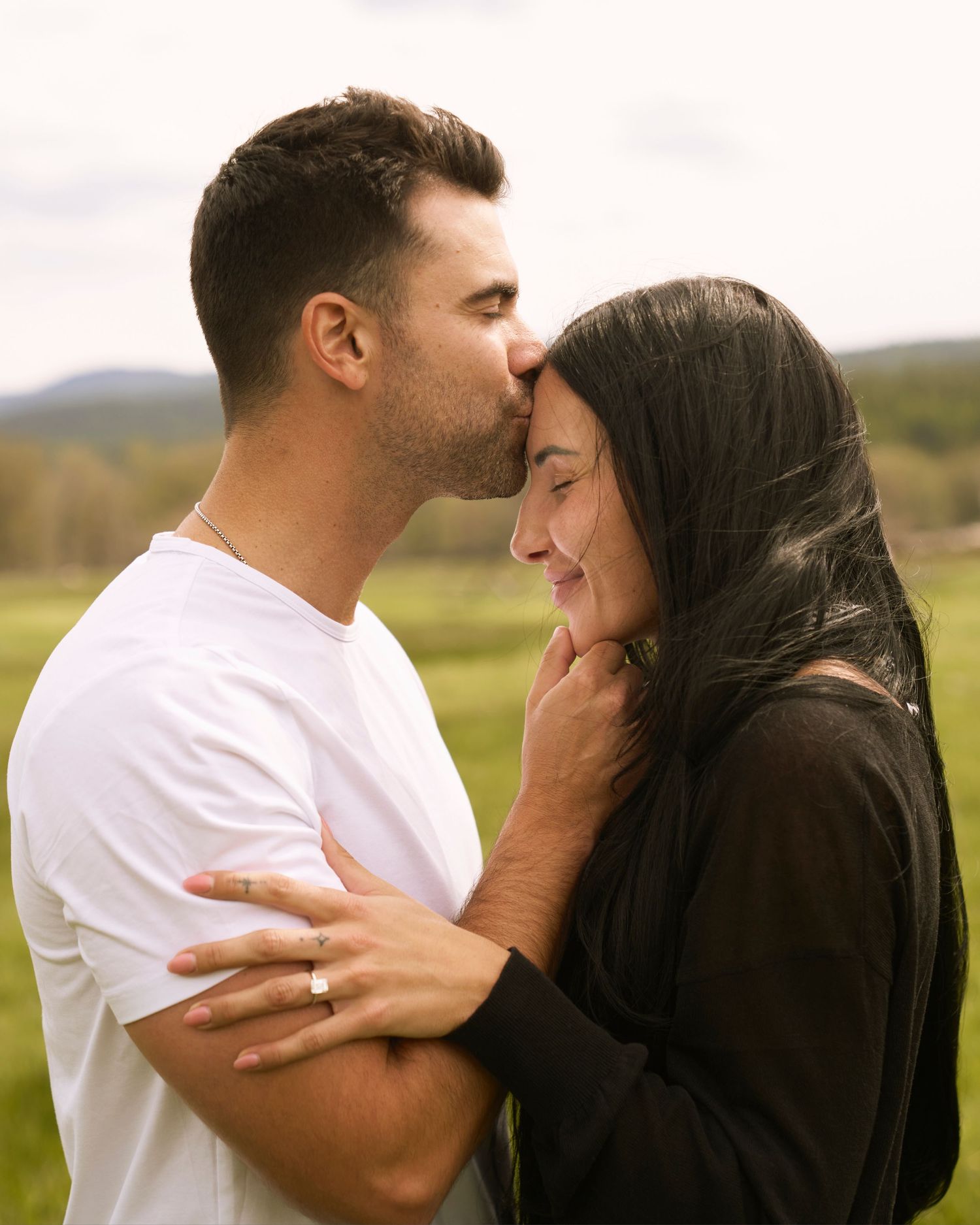 Man in white tee kissing womans forehead in green field.