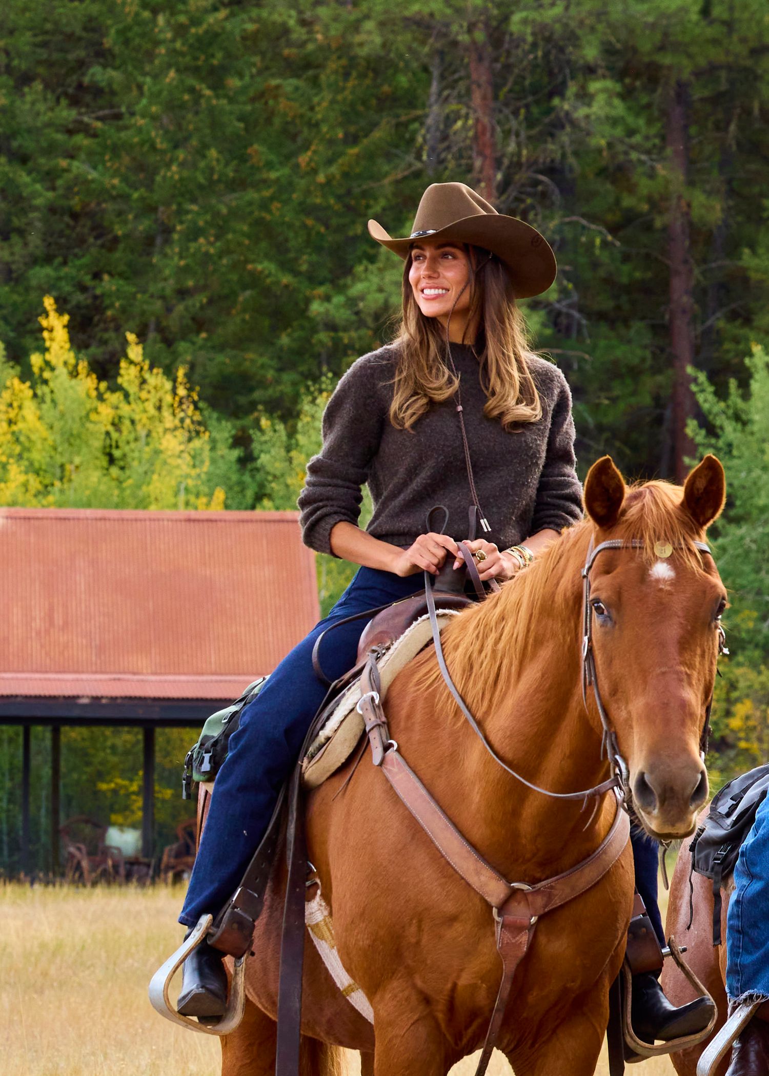 Smiling woman in brown cowboy hat riding chestnut horse in outdoor ranch setting.