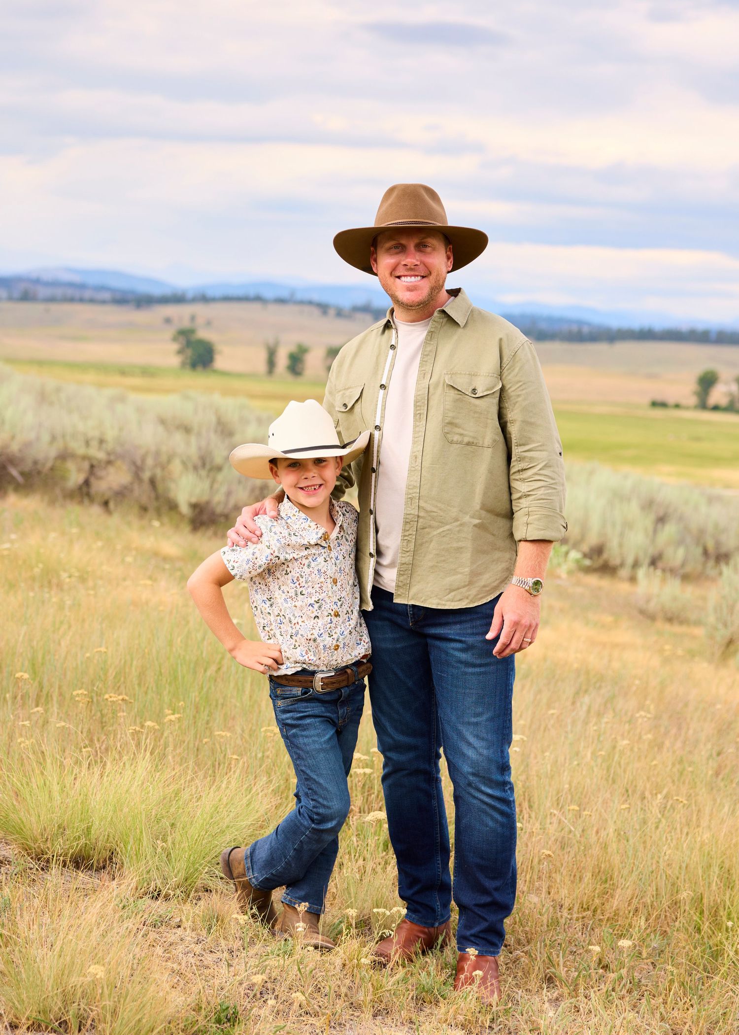 Smiling father and son in cowboy hats posing together in a wide open sagebrush prairie landscape.