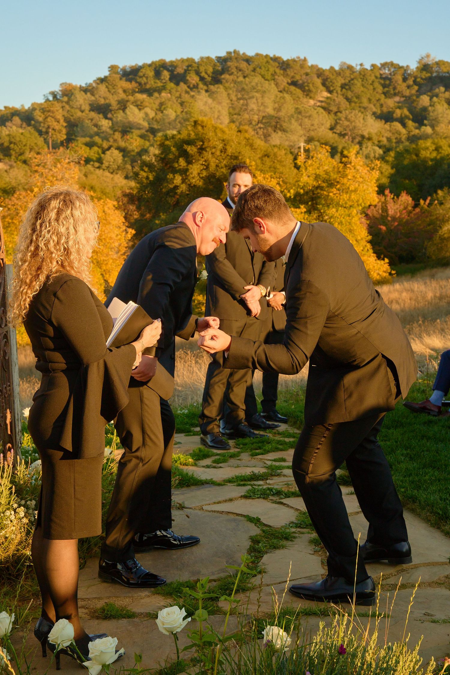 Groomsmen in dark suits share a joyful moment during outdoor wedding ceremony with autumn hillside backdrop.