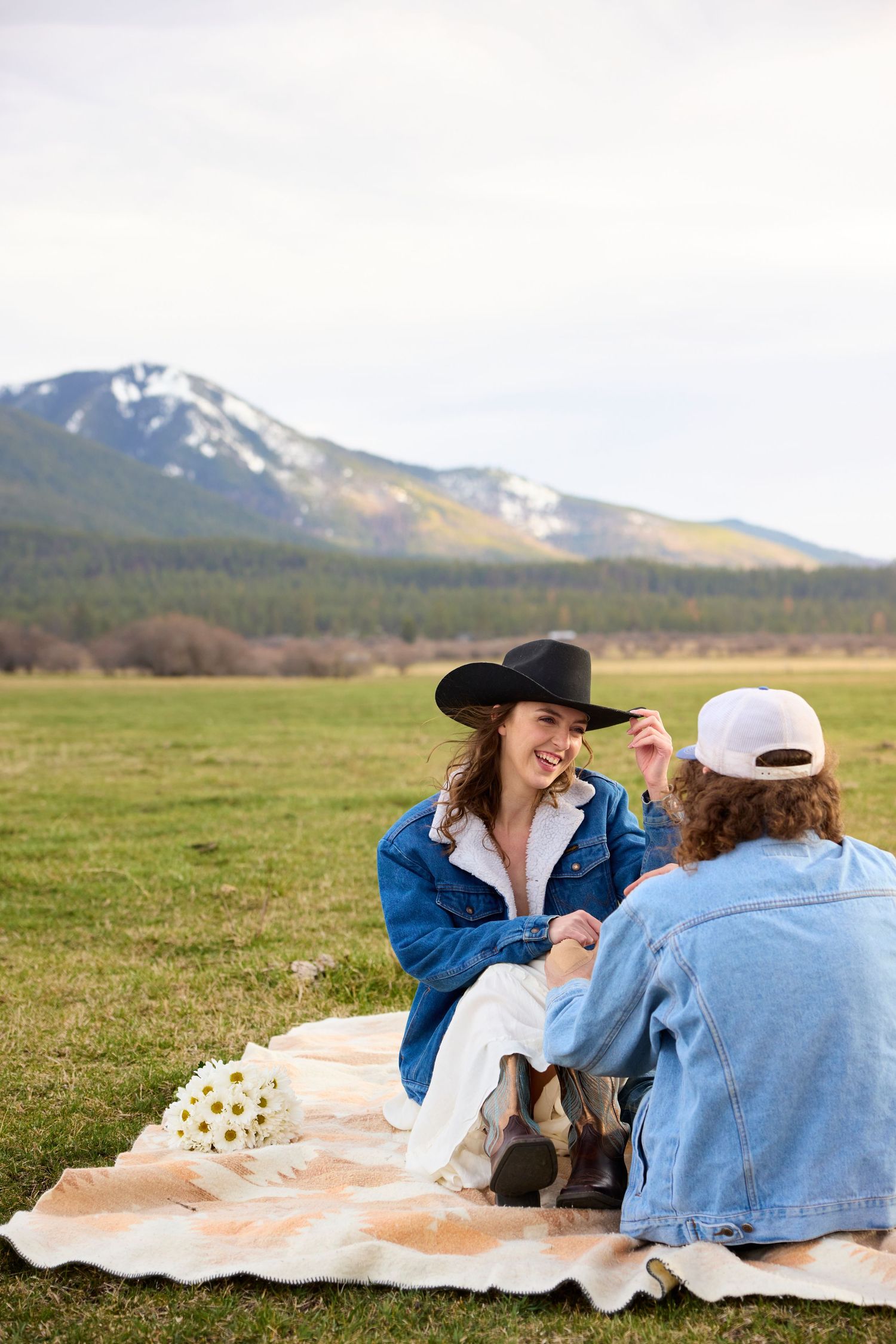 Couple sitting on a blanket in a mountain meadow, woman in black cowboy hat laughing with bouquet of daisies.