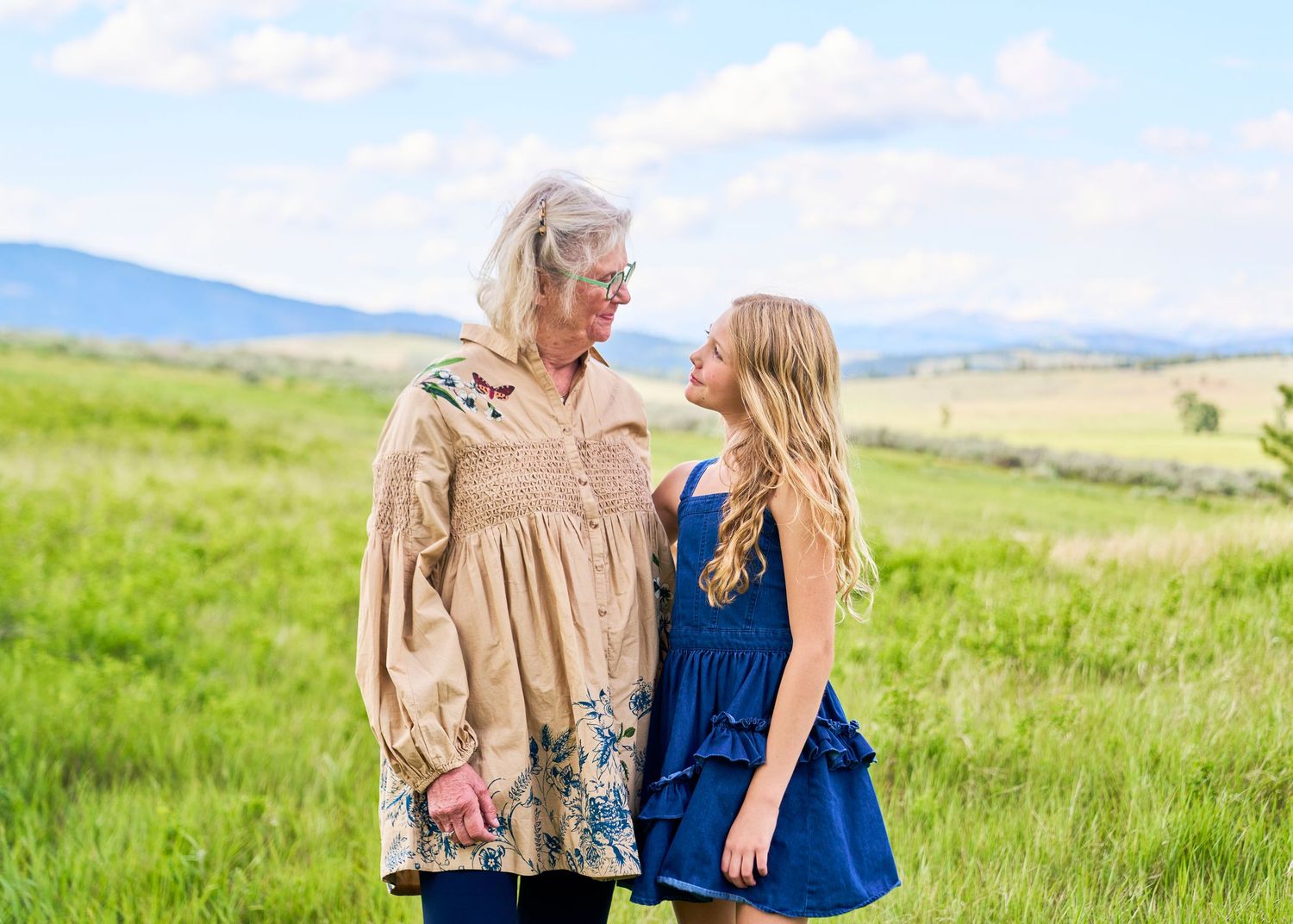 Grandmother and granddaughter sharing a tender moment in a green field with mountains in the background.