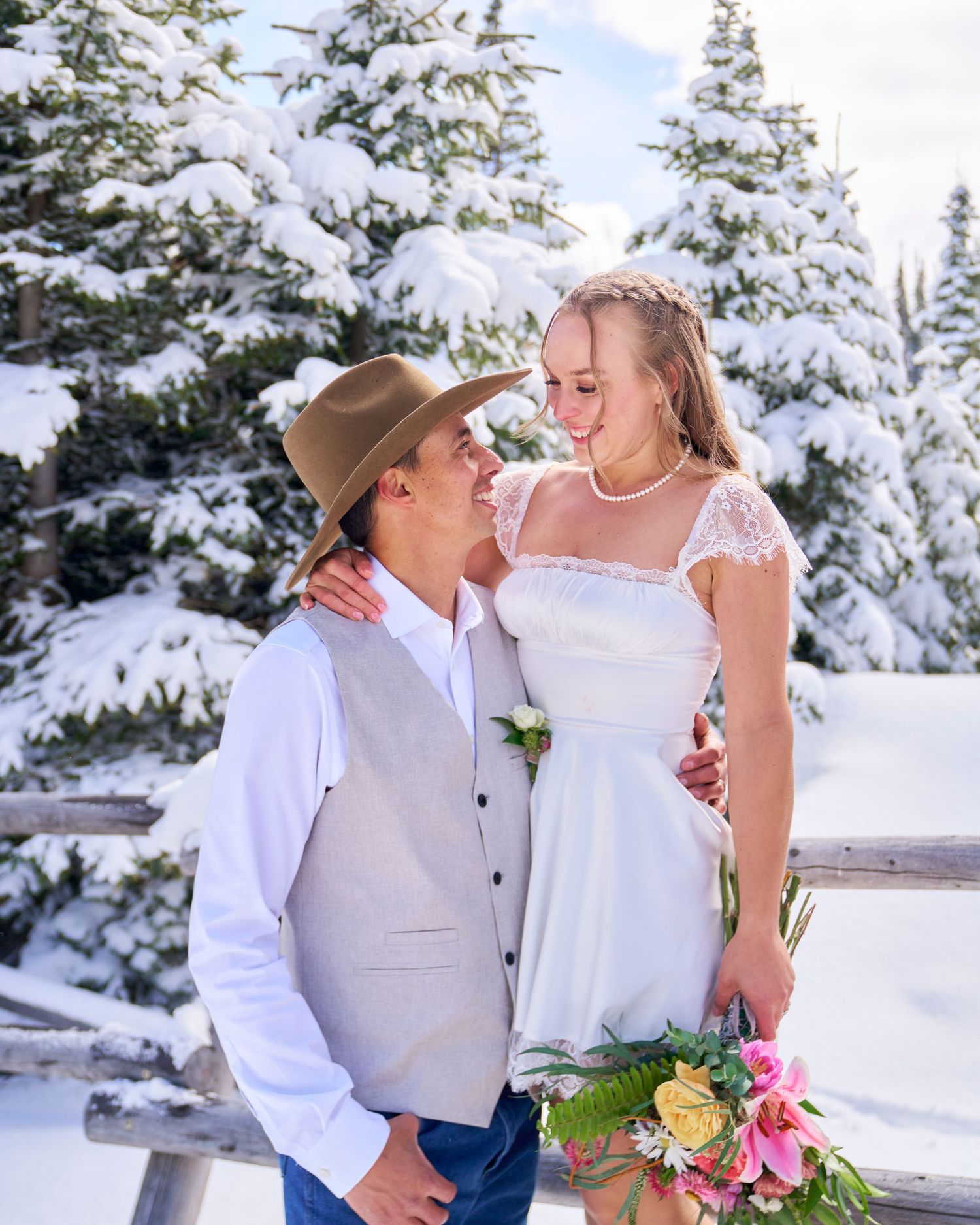 Wedding couple smiling at each other by rustic fence, bride holding colorful bouquet in winter snow.