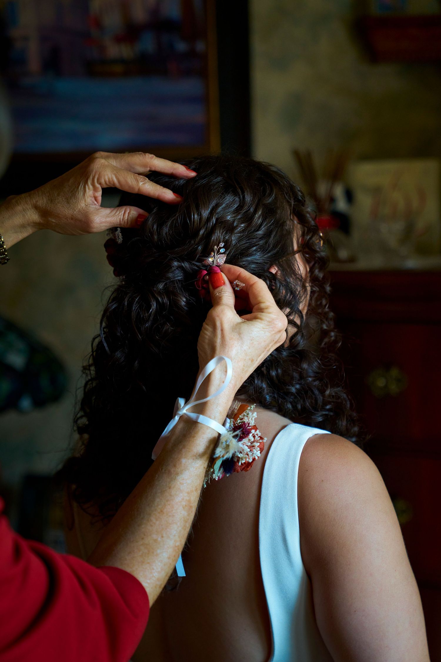 Bride with dark curly hair getting red floral hair accessory carefully placed during pre-wedding preparations indoors.