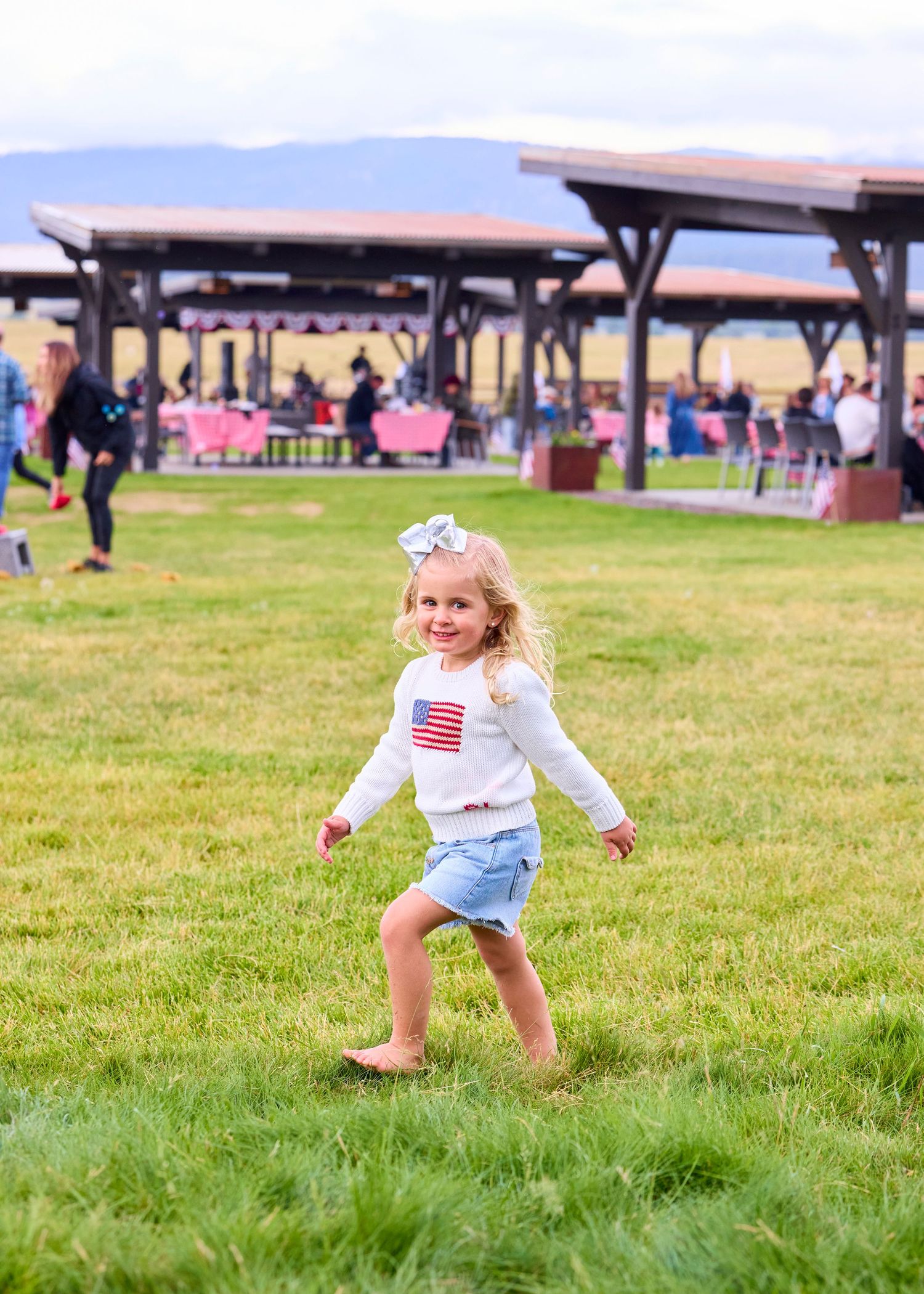 Smiling young girl in American flag shirt running barefoot on green grass at outdoor event.