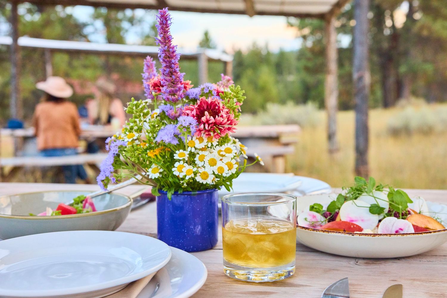 Rustic outdoor dining table with wildflower bouquet, whiskey cocktail, and fresh salad in natural setting.