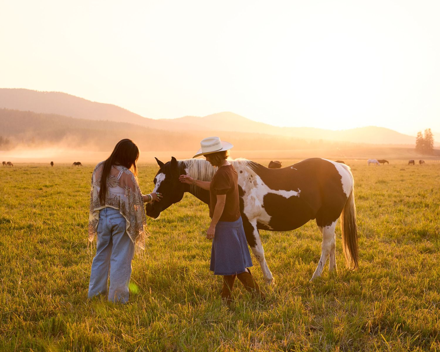 Two women interact with a black and white paint horse in a golden sunset field with mountains in the background.