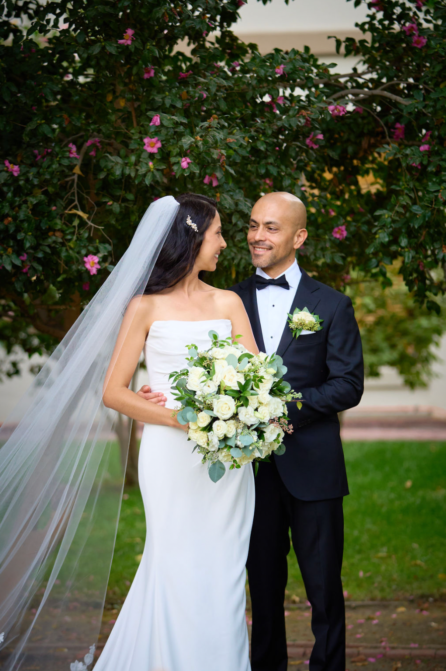 Elegant bride and groom smiling at each other, she holds white bouquet, blooming tree backdrop.