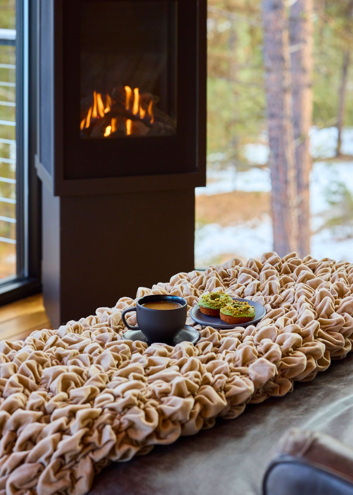 A cozy interior scene with a glowing fireplace, textured rug, coffee cup, and colorful cupcakes beside snowy windows.