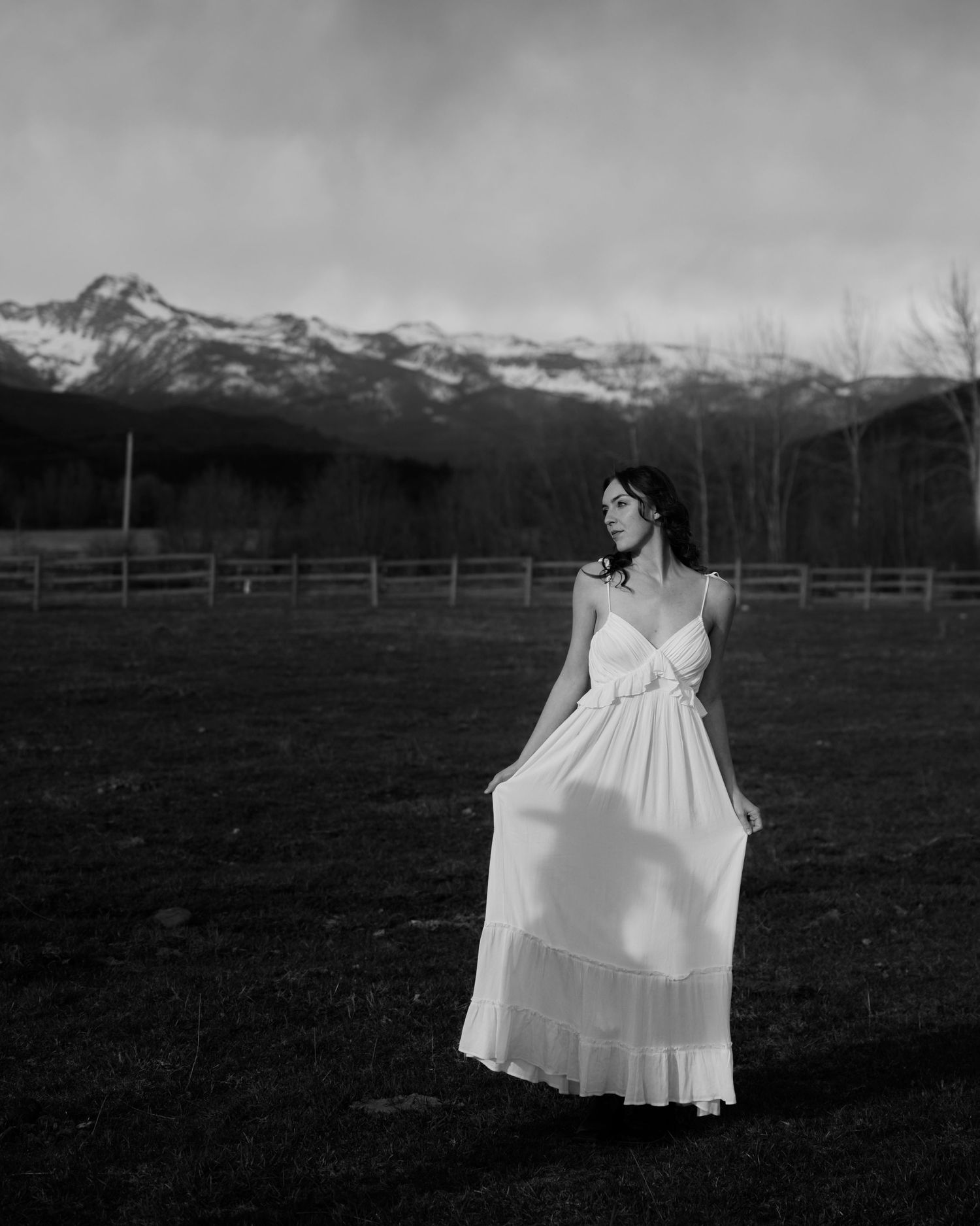 Woman in white dress stands in field with snow-capped mountains behind her in black and white.