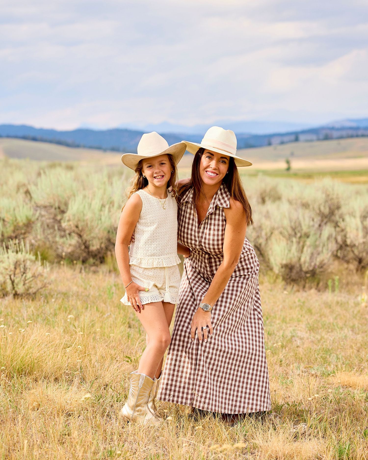 Mother and daughter in coordinating western outfits and cowgirl hats posing in sage brush field.