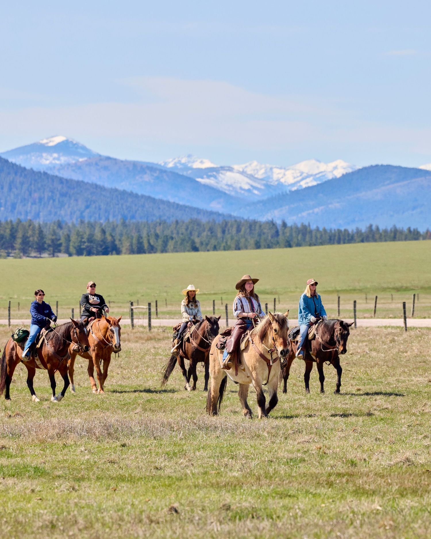 Group of horseback riders crossing green meadows with snow-capped mountains behind.