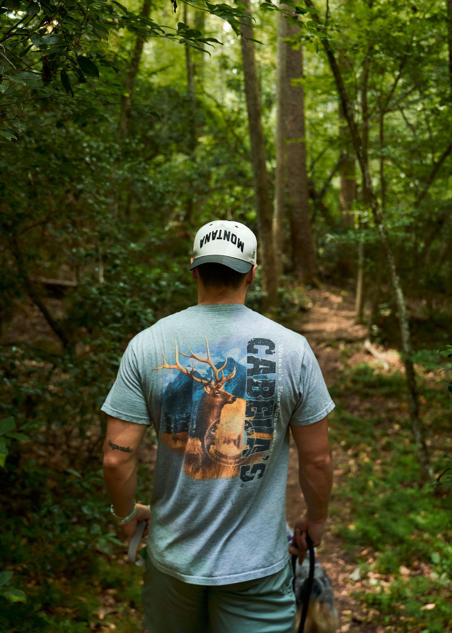 Man in Cabela's t-shirt and Montana cap hiking through lush green forest trail.