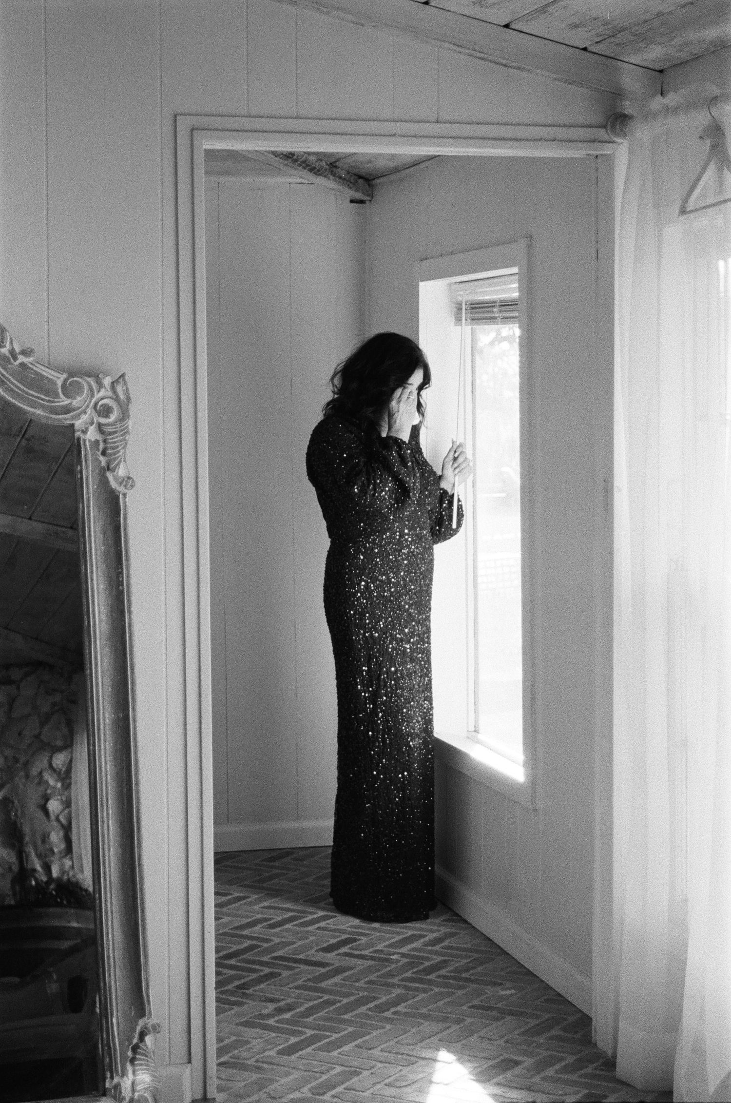Elegant woman in black sequin gown stands by window in black and white film photography portrait.