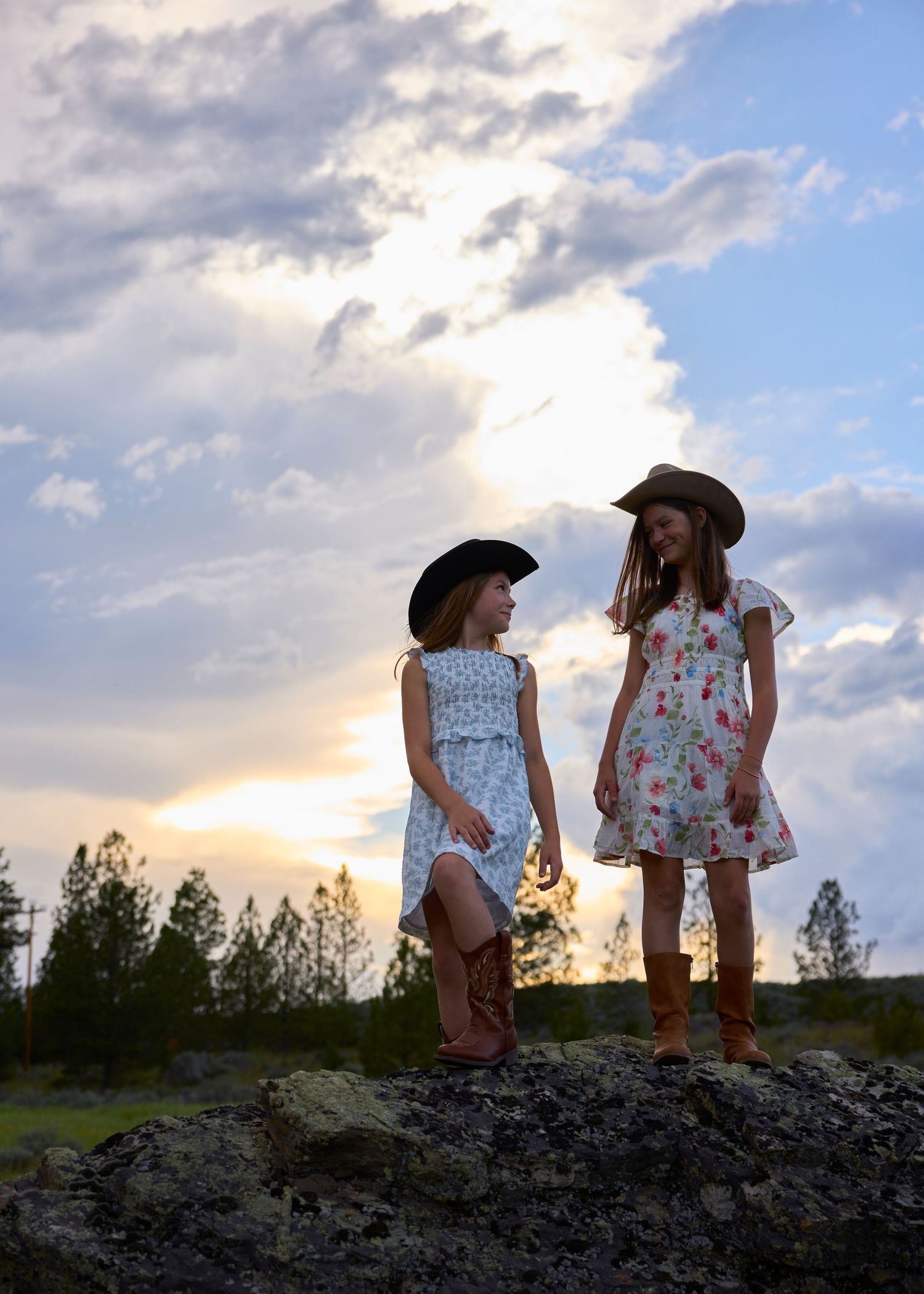 Two girls in cowboy hats and boots stand on a rock at sunset in a rural landscape.