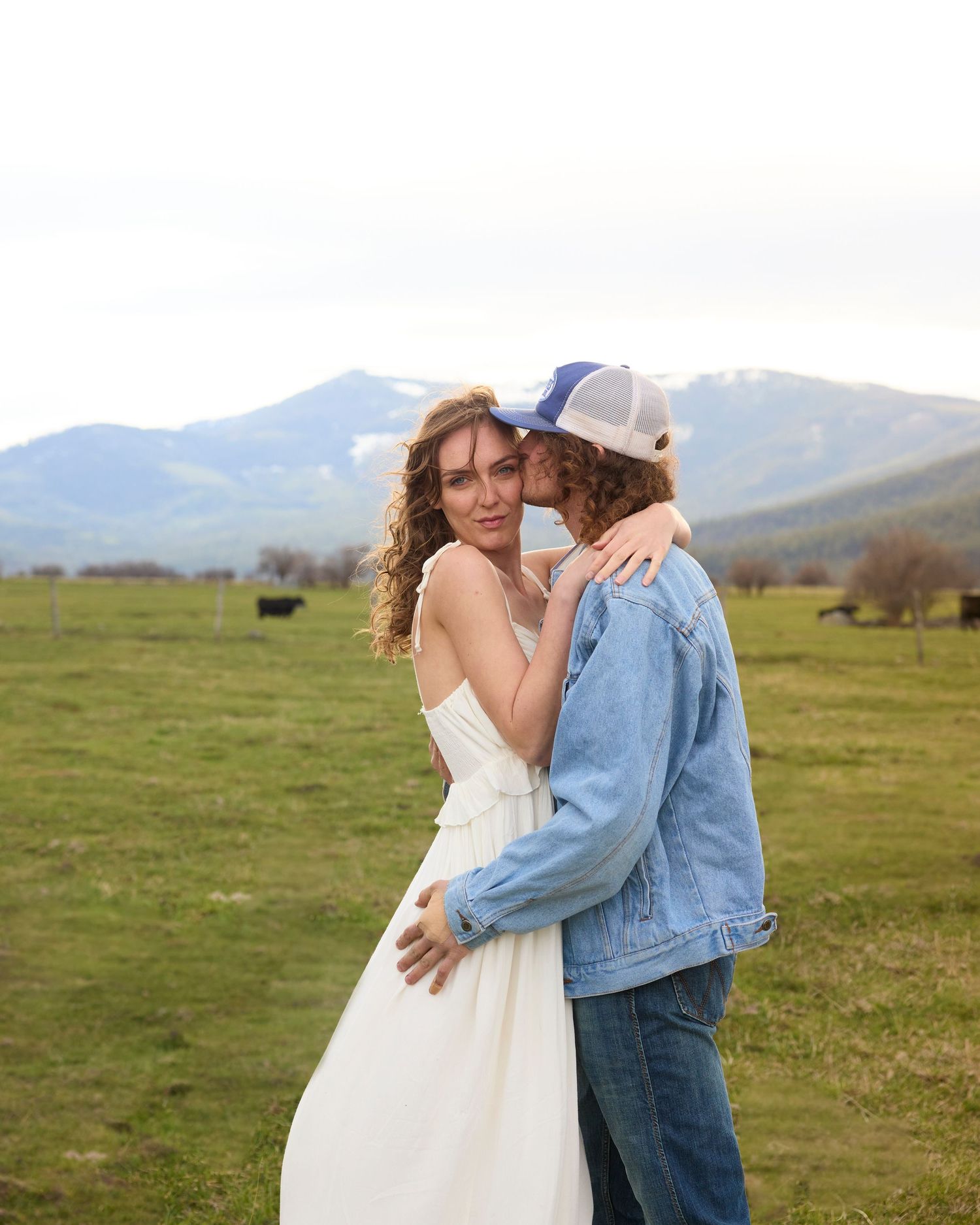 A couple embracing in a green field with mountain backdrop, woman in white dress.