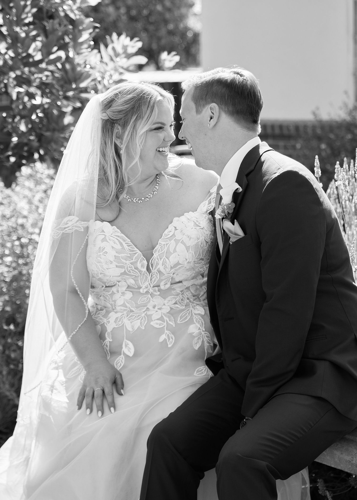 Black and white photo of smiling bride in lace gown and groom sitting together outdoors.