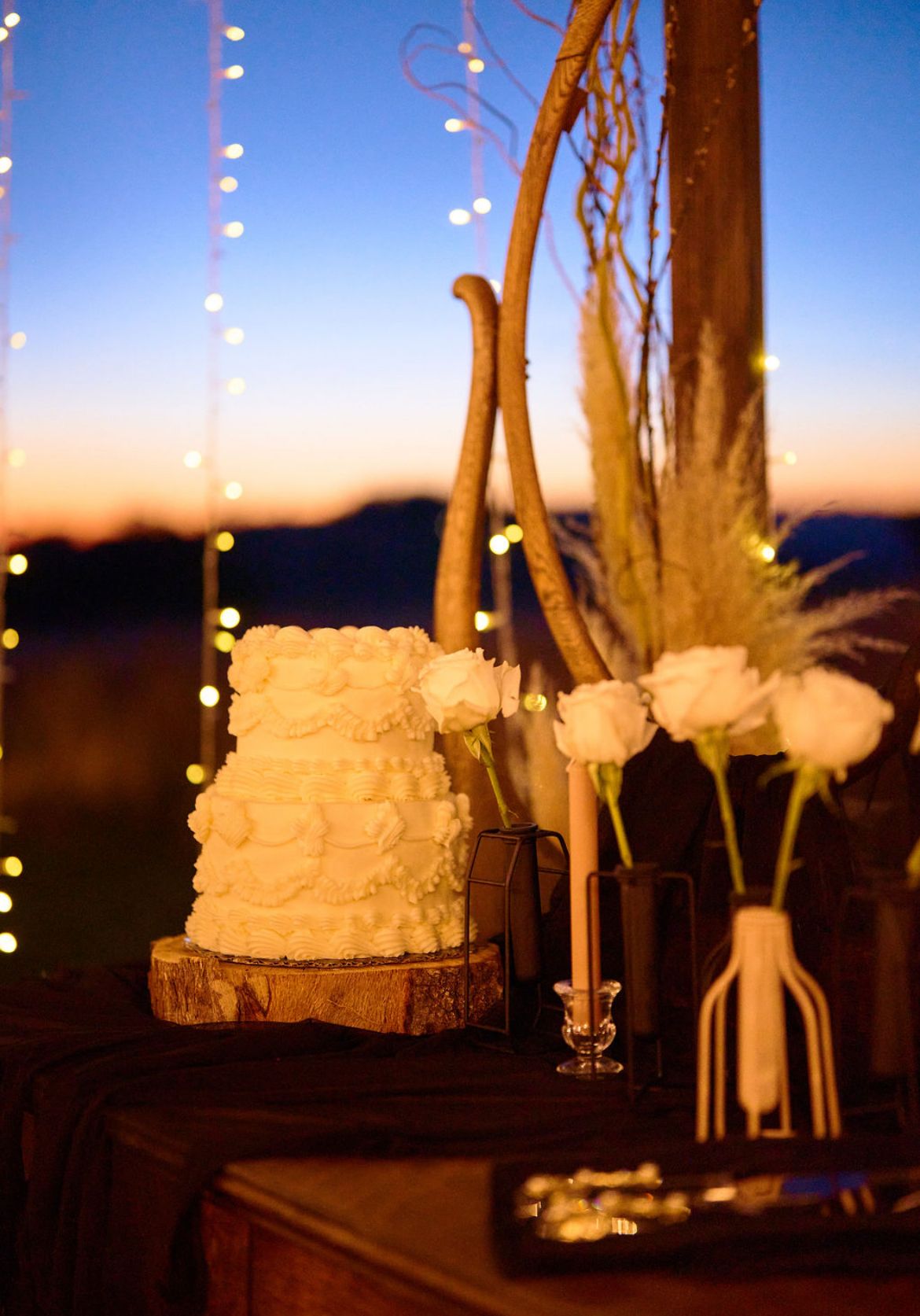 Elegant white wedding cake on wood slice with white roses and fairy lights at twilight outdoor reception.
