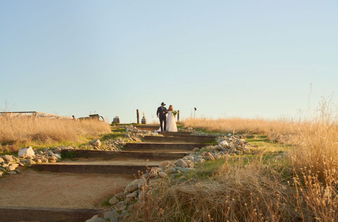 Bride and groom stand together on stone steps surrounded by golden grass at outdoor wedding venue.