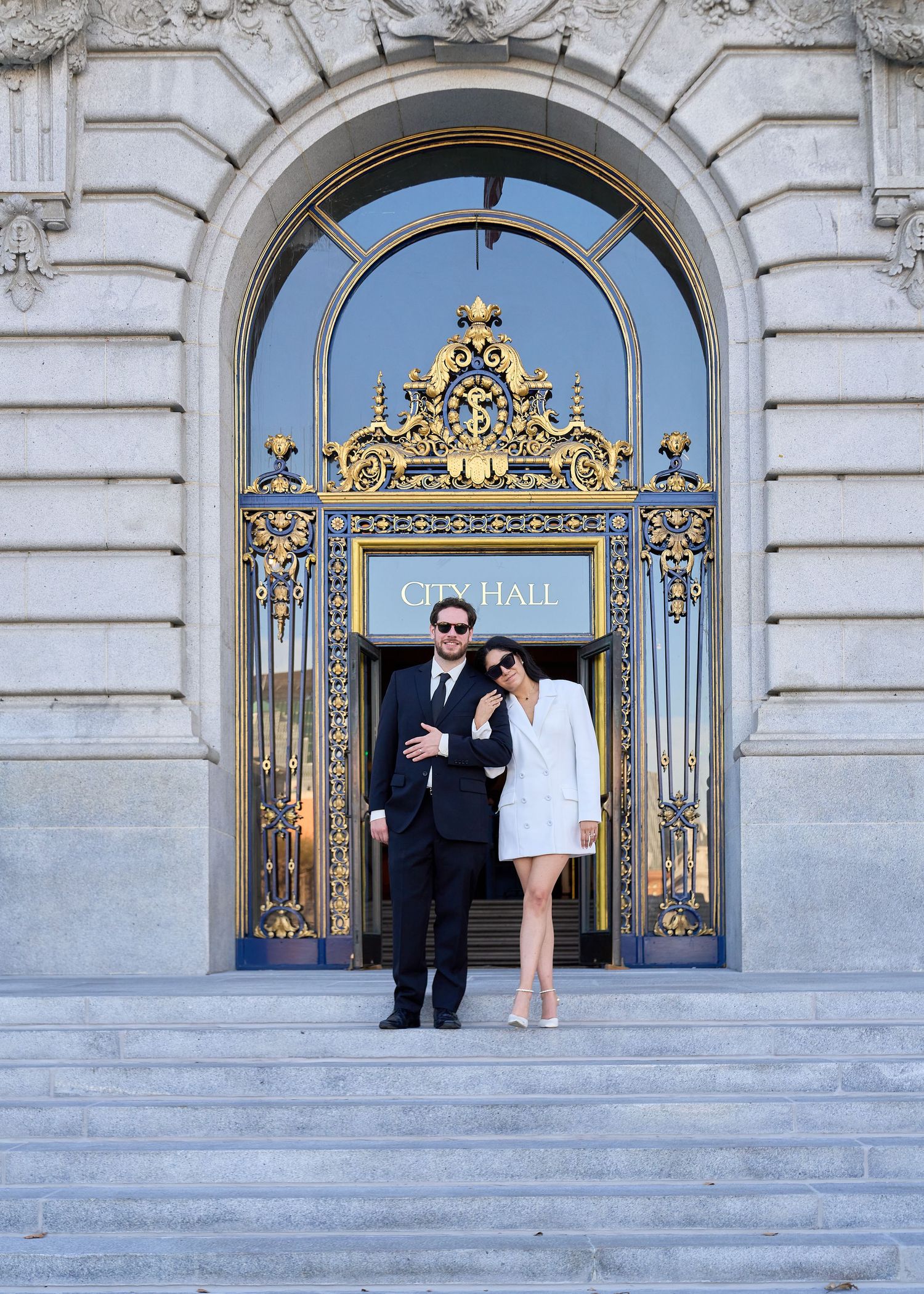Newlyweds pose in front of ornate gold City Hall entrance gates, bride in white blazer dress.