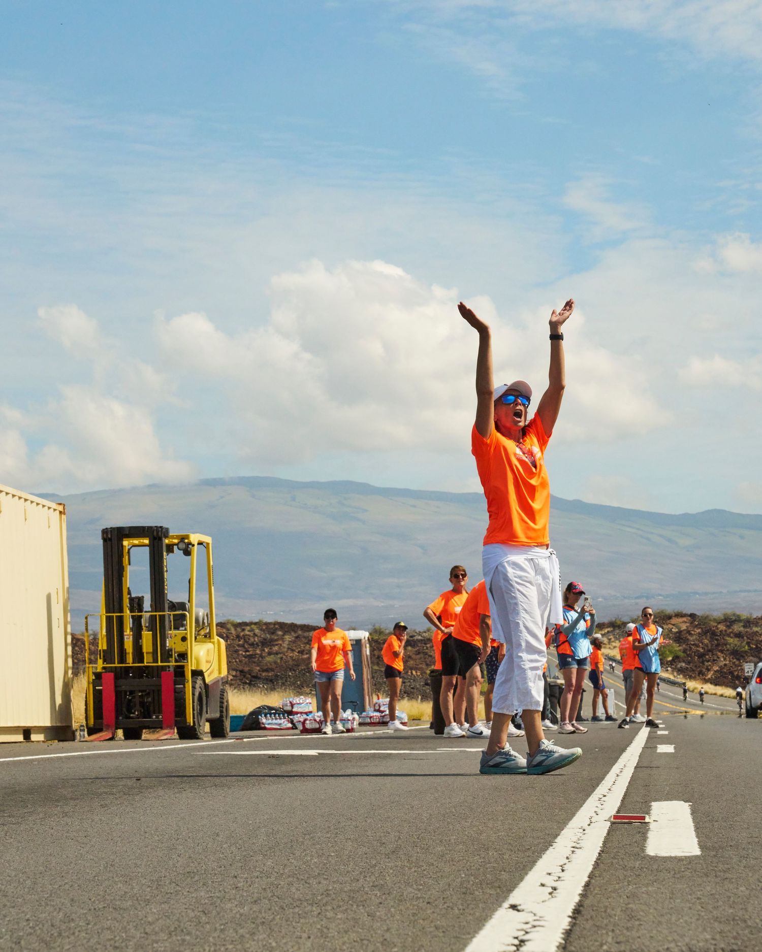 Person in orange shirt raises hands on road with forklift and mountains in background during outdoor event.