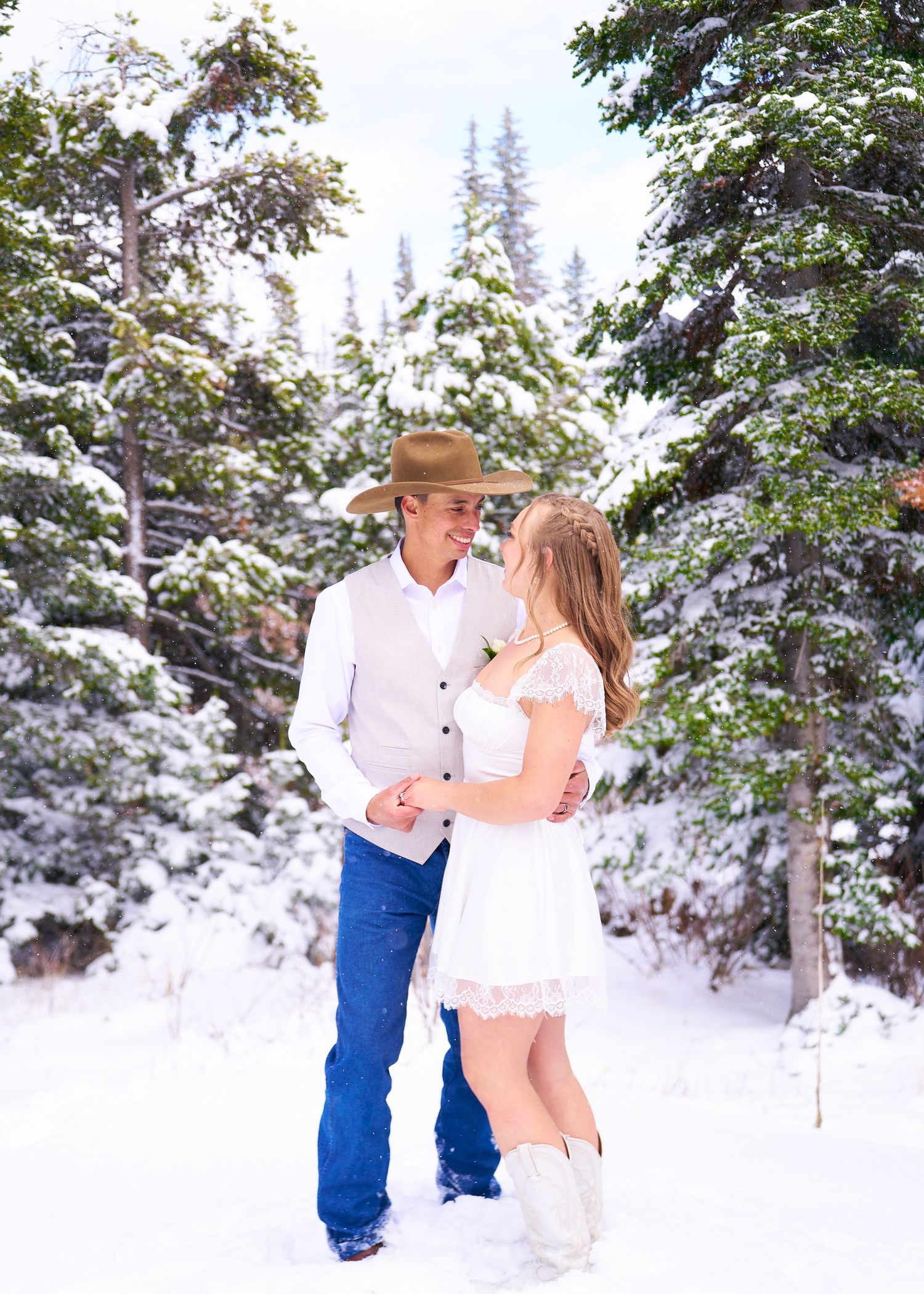 Groom in cowboy hat and white vest smiles at bride in white lace dress in snowy pine forest.