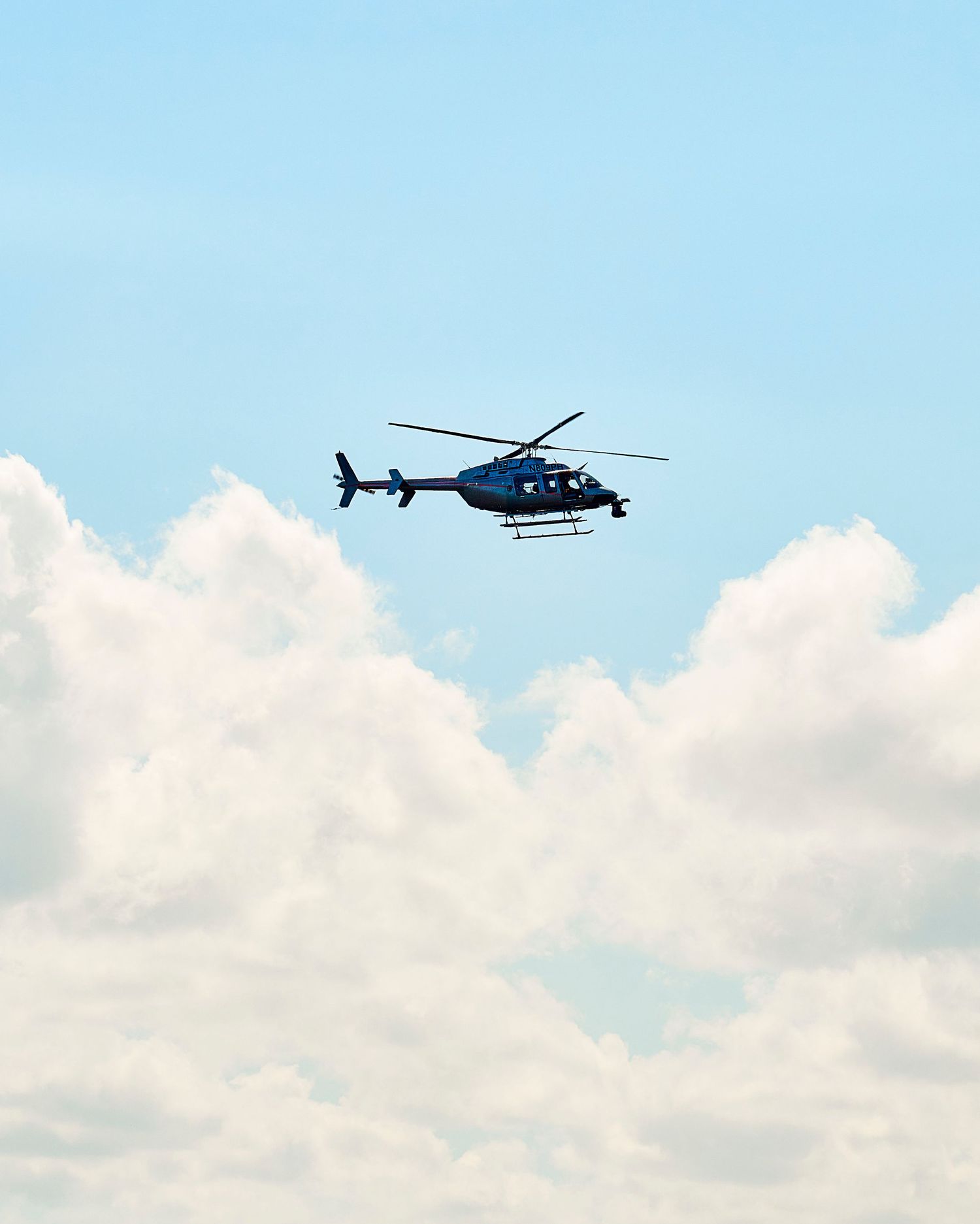 A blue helicopter flying through a partly cloudy sky with white cumulus clouds below.