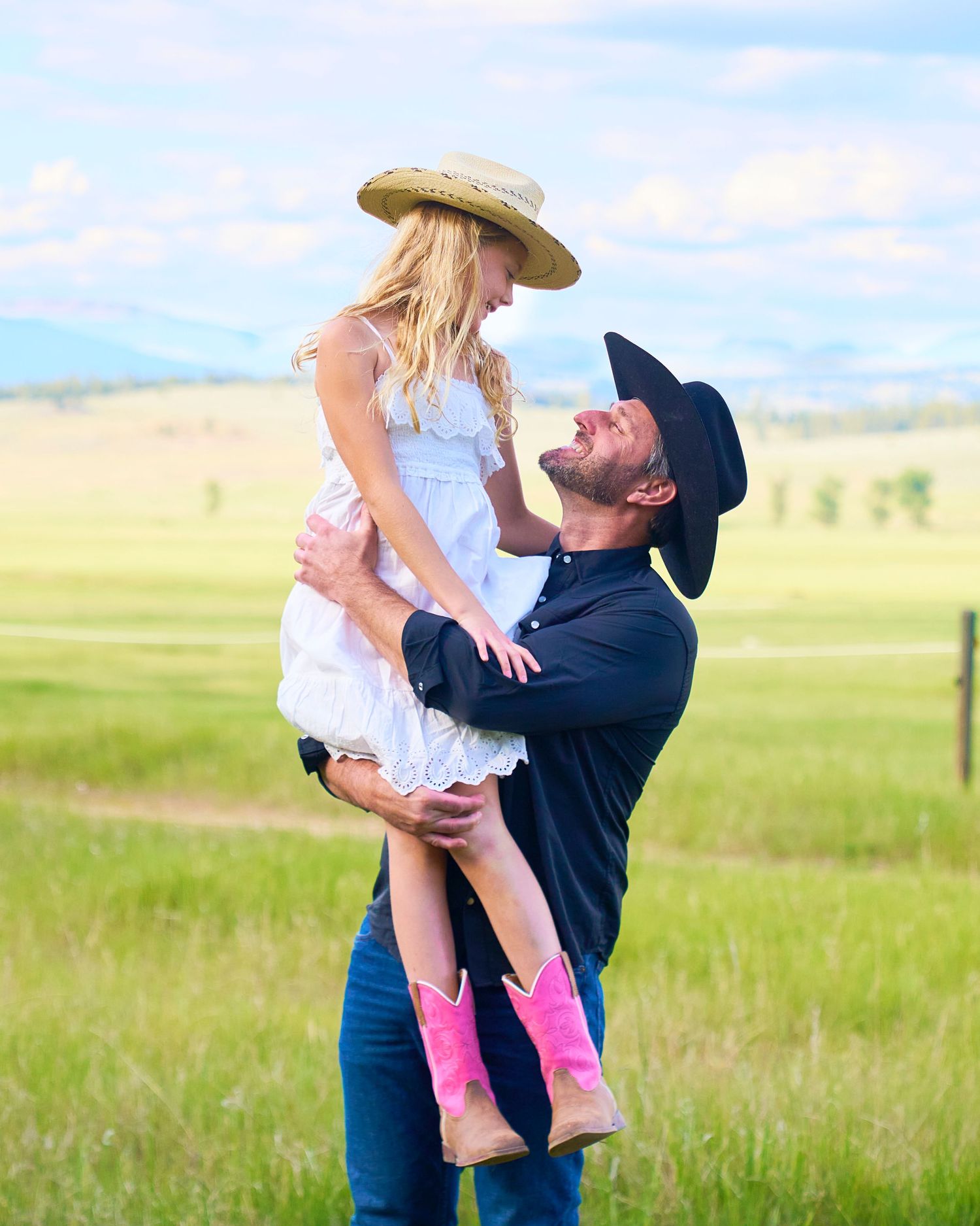 Man in black cowboy hat lifts smiling girl in white dress and pink boots in green meadow.