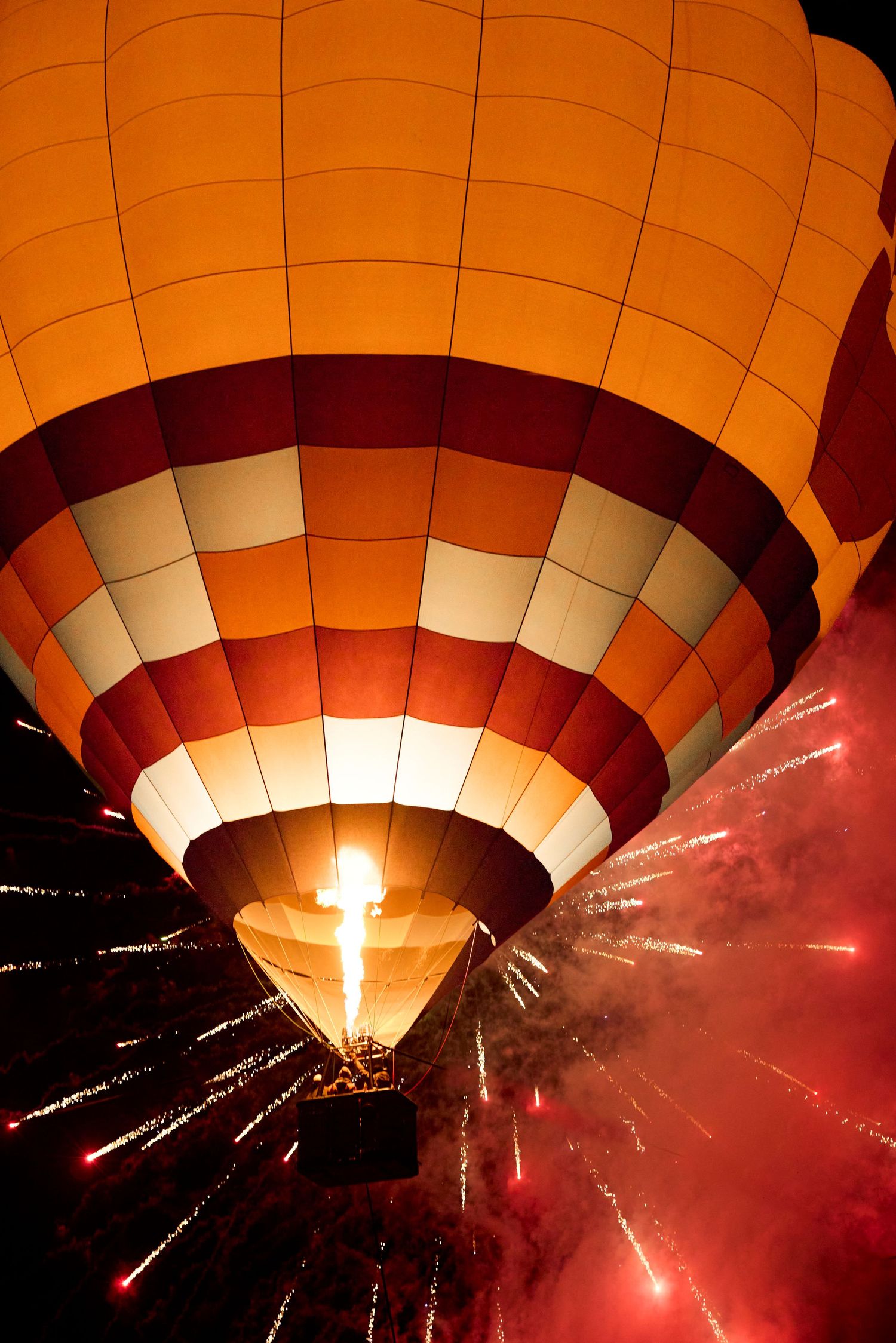 A glowing orange hot air balloon illuminated at night with colorful fireworks exploding nearby.