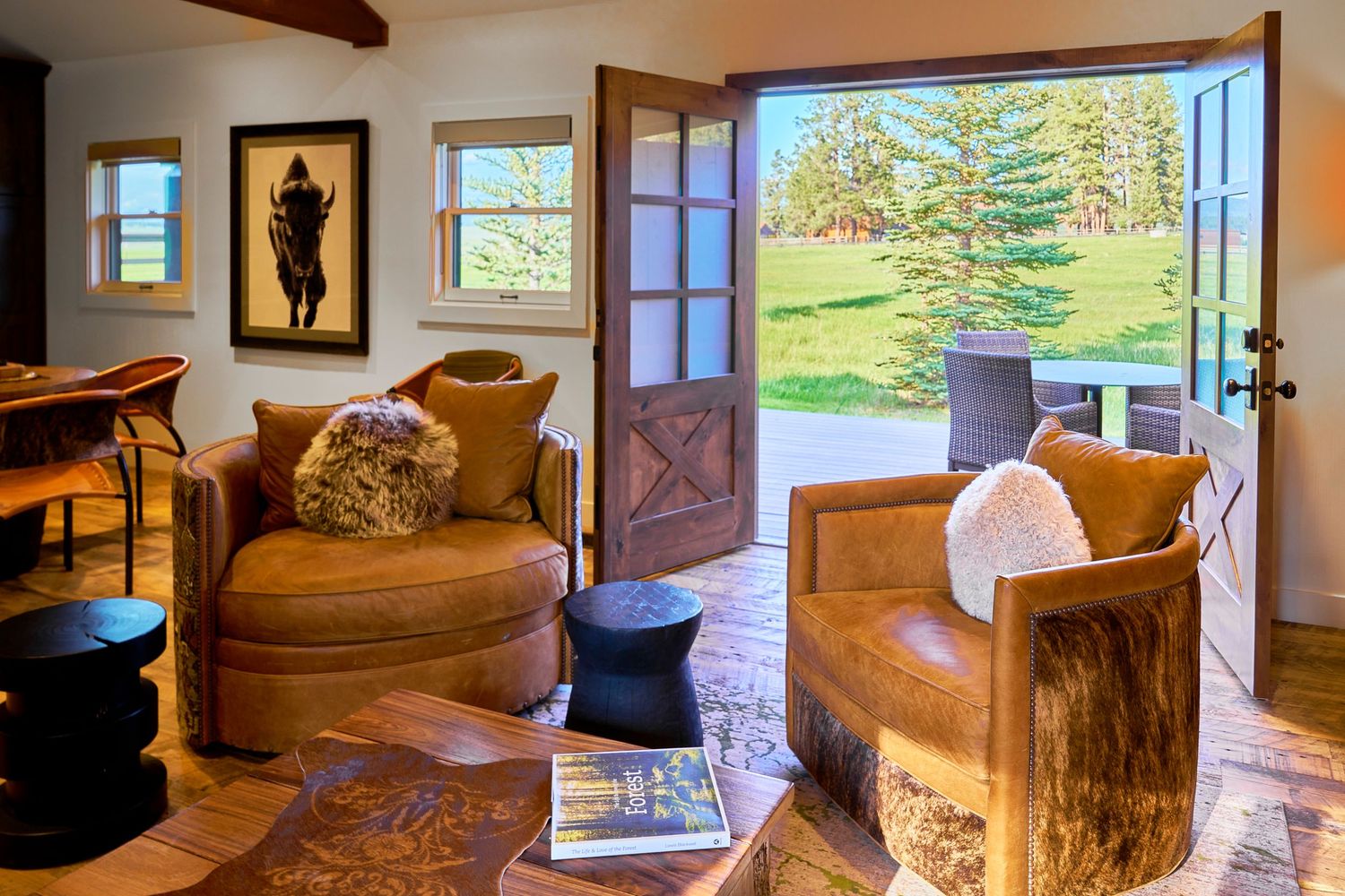 Cozy rustic lodge interior with leather armchairs, open French doors overlooking green meadow landscape.