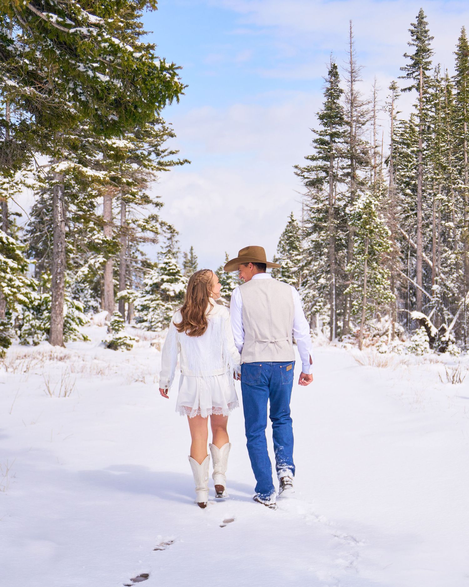 Bride in white dress and cowboy boots walks with groom in cowboy hat along snowy forest trail.