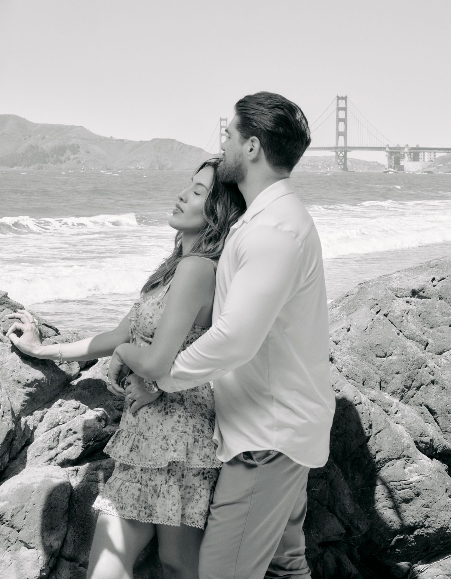 Black and white photo of a couple embracing on rocky shore with Golden Gate Bridge in background.