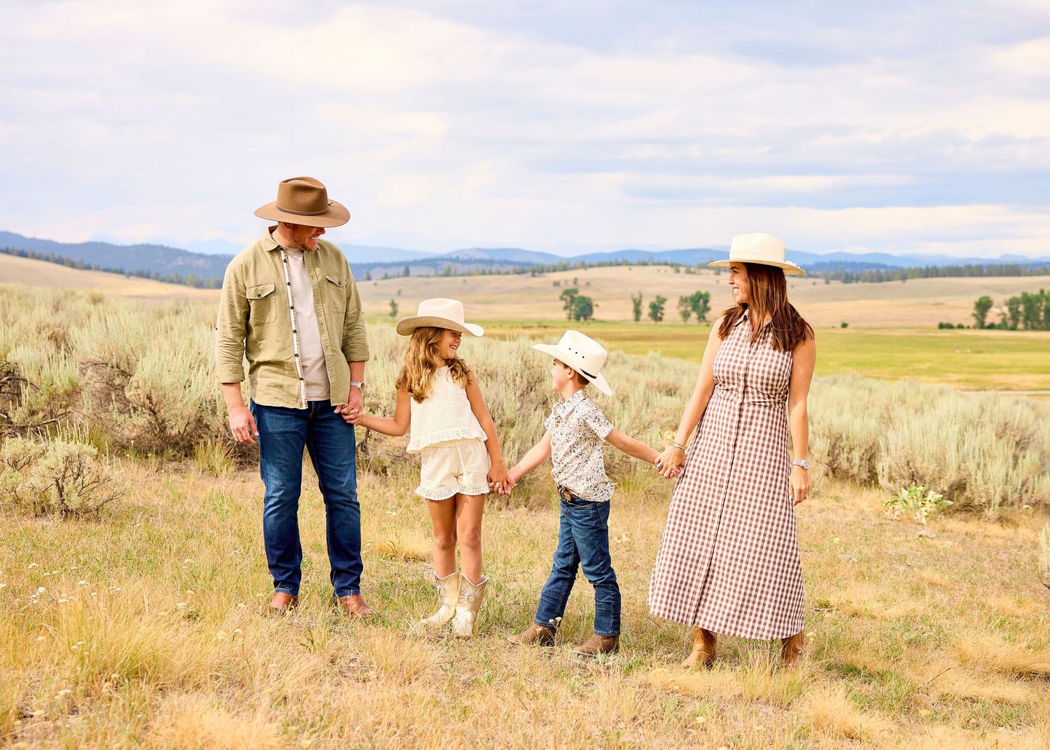 Family of four in western attire holding hands in golden sagebrush meadow with mountains.